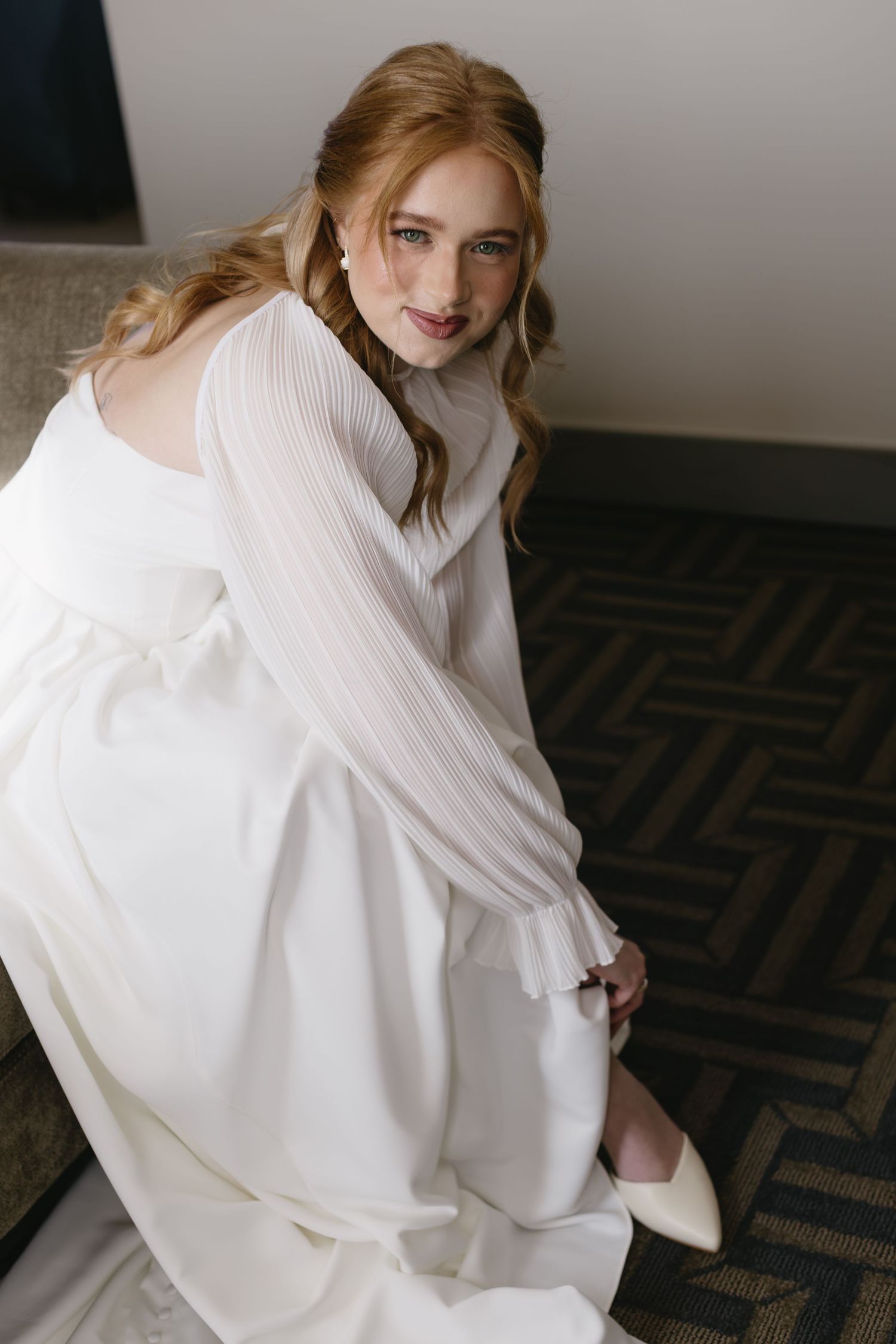 Bride in an elegant white long-sleeved wedding gown sits gracefully on a patterned carpet.