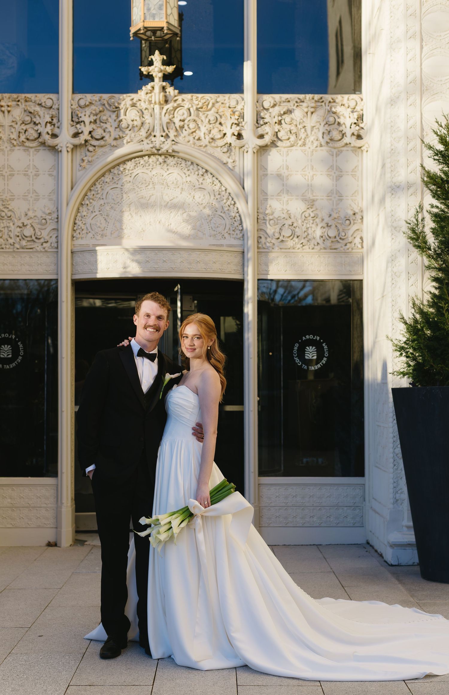 Elegant wedding couple poses against ornate white architectural backdrop with flowing bridal train.
