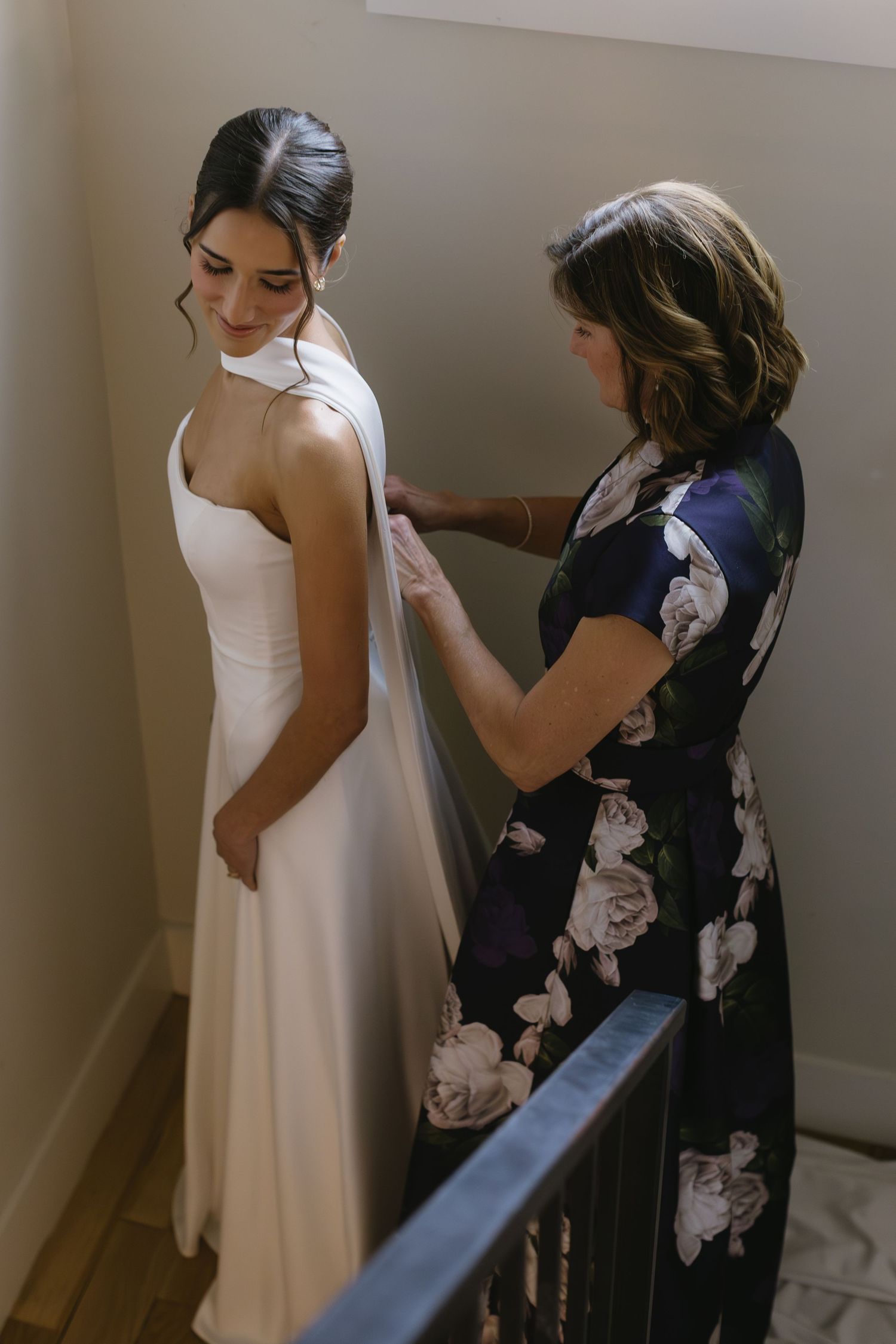 A bride in a sleek white gown gets help with final adjustments from someone in a floral dress.
