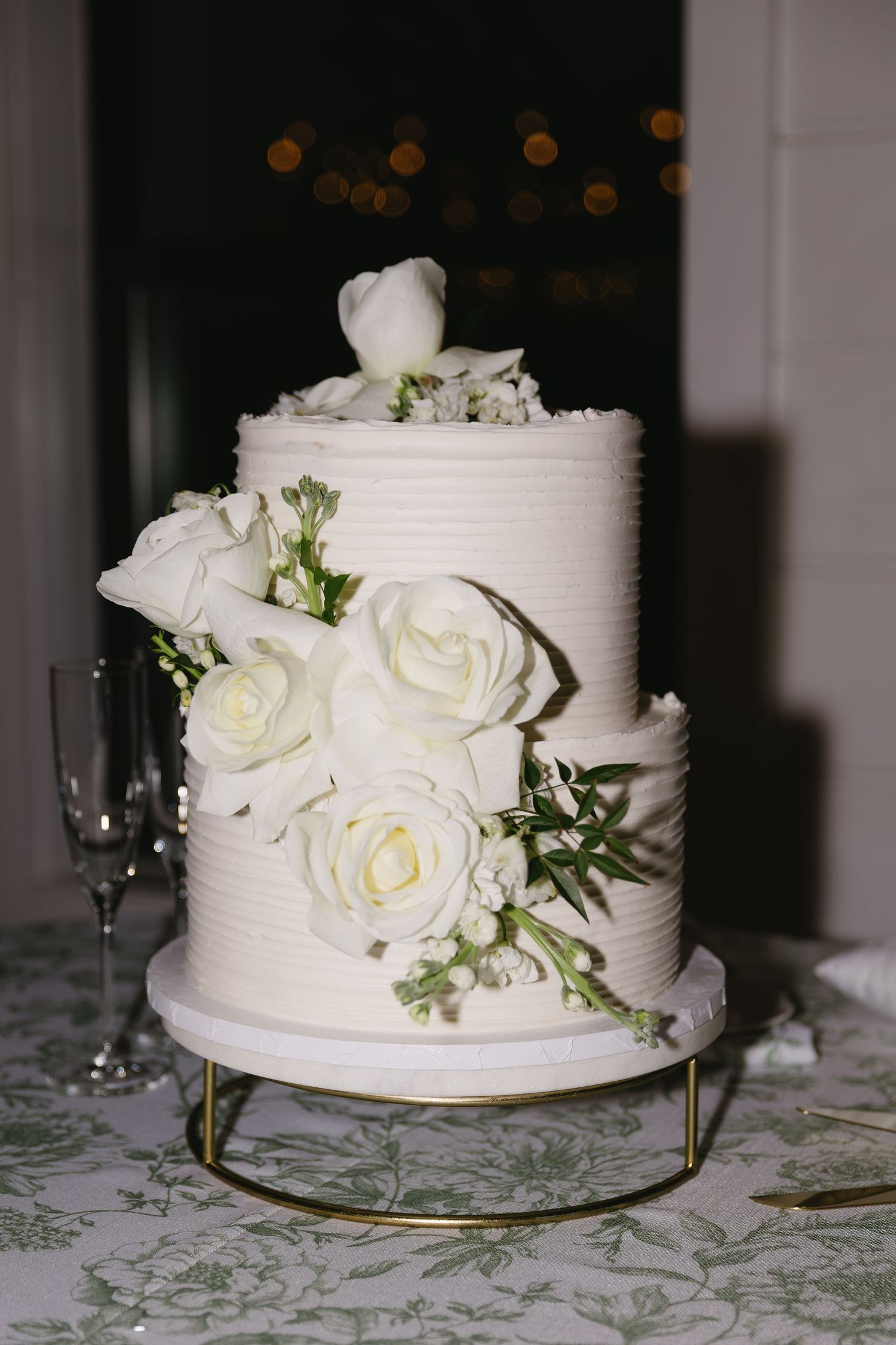 Classic white wedding cake decorated with fresh roses and greenery on gold stand.