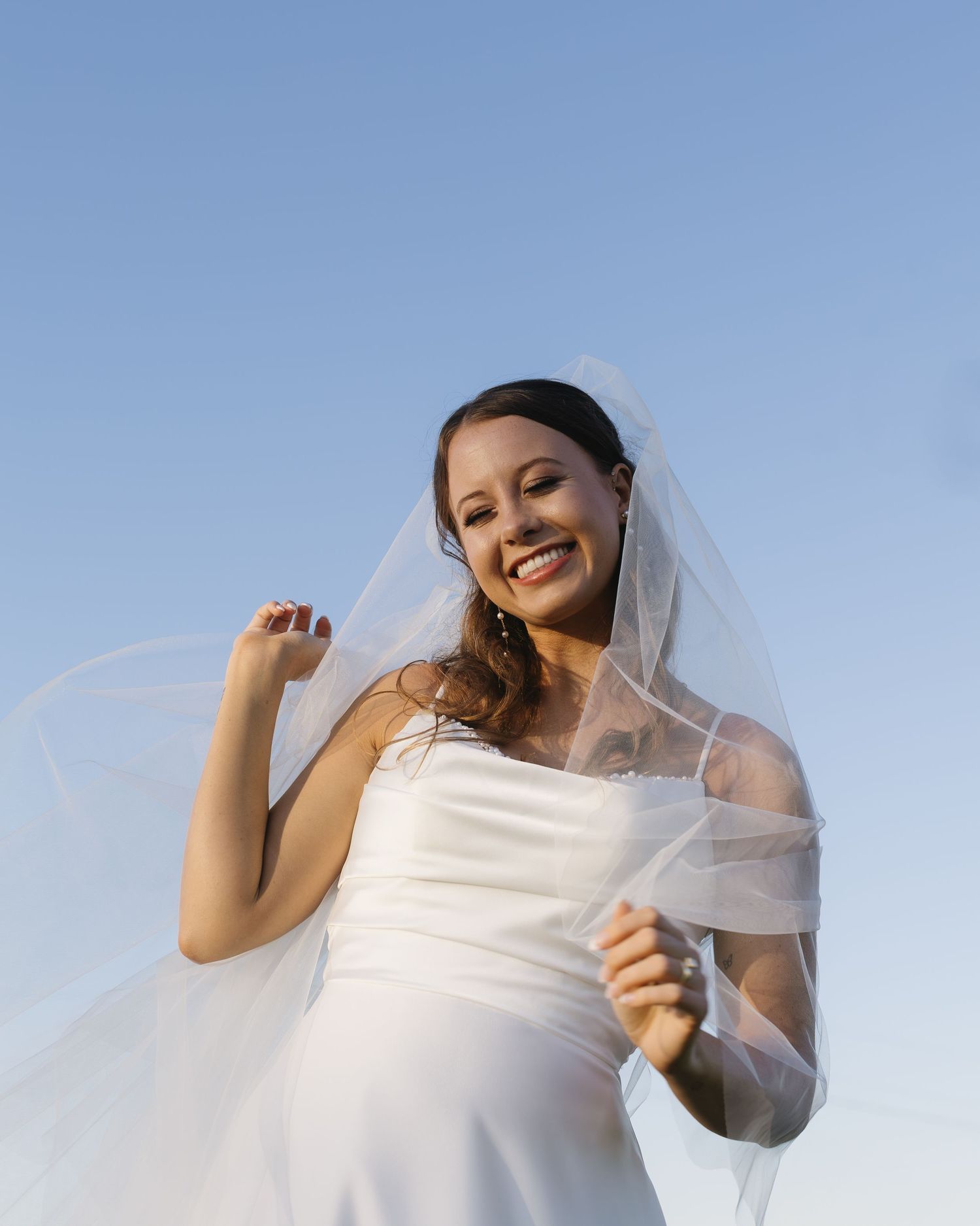 Candid bridal portraits against blue sky showing movement and joy with flowing veil.
