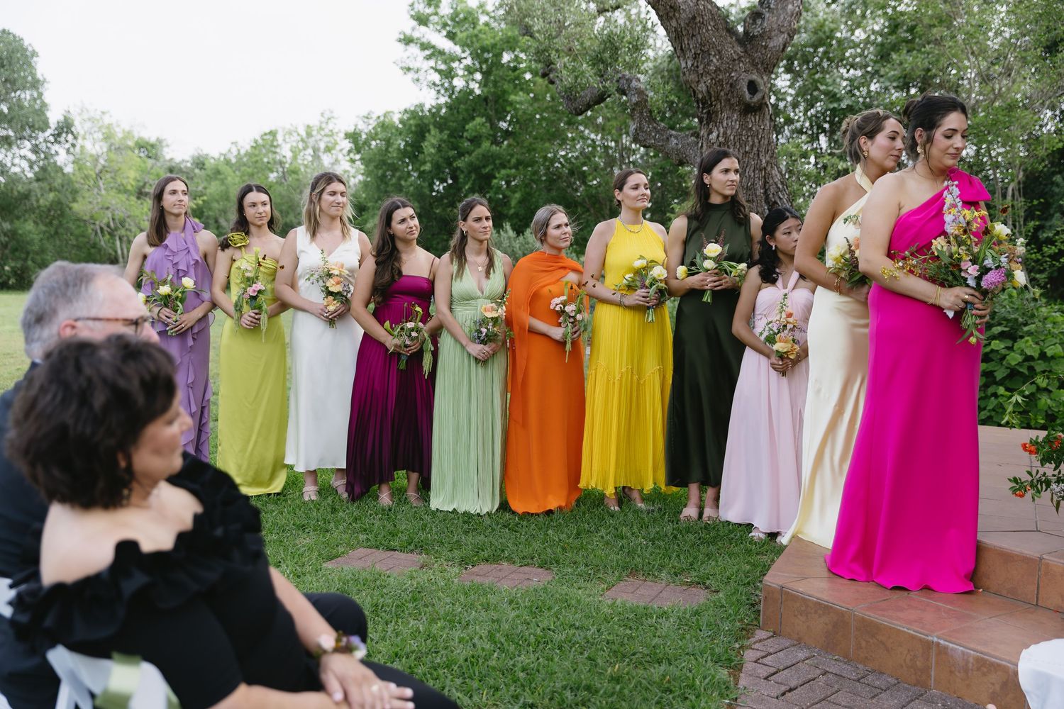 Row of bridesmaids in colorful rainbow dresses standing on grass at outdoor wedding.