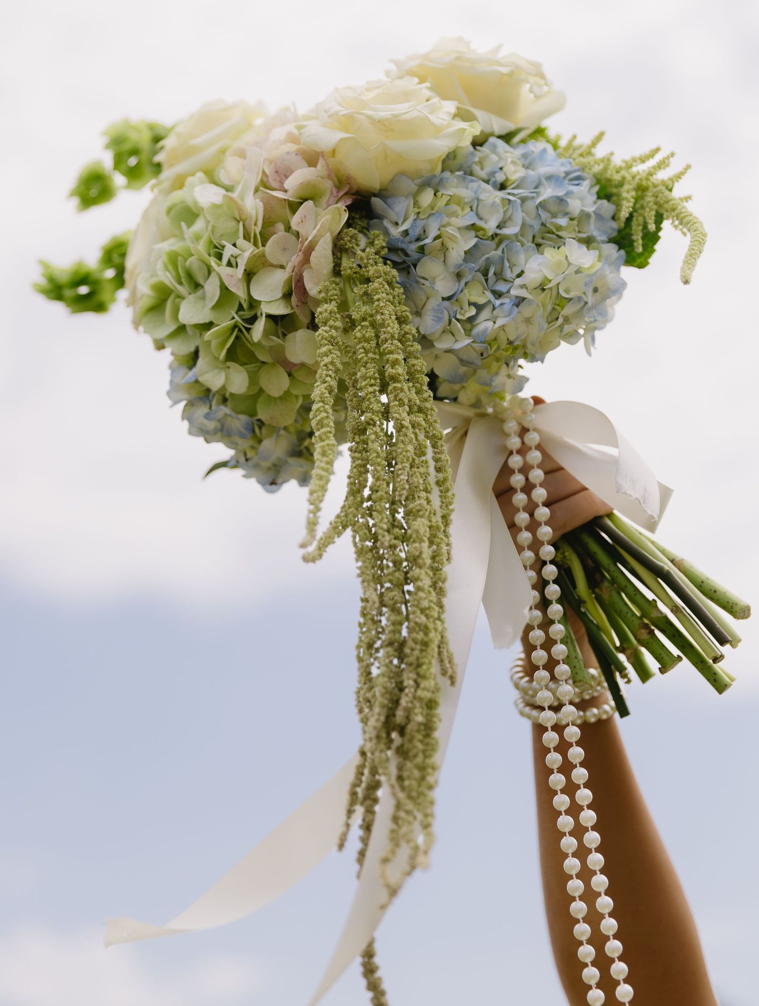 Wedding bouquet with cascading white flowers and pearl strands held against a bright sky.