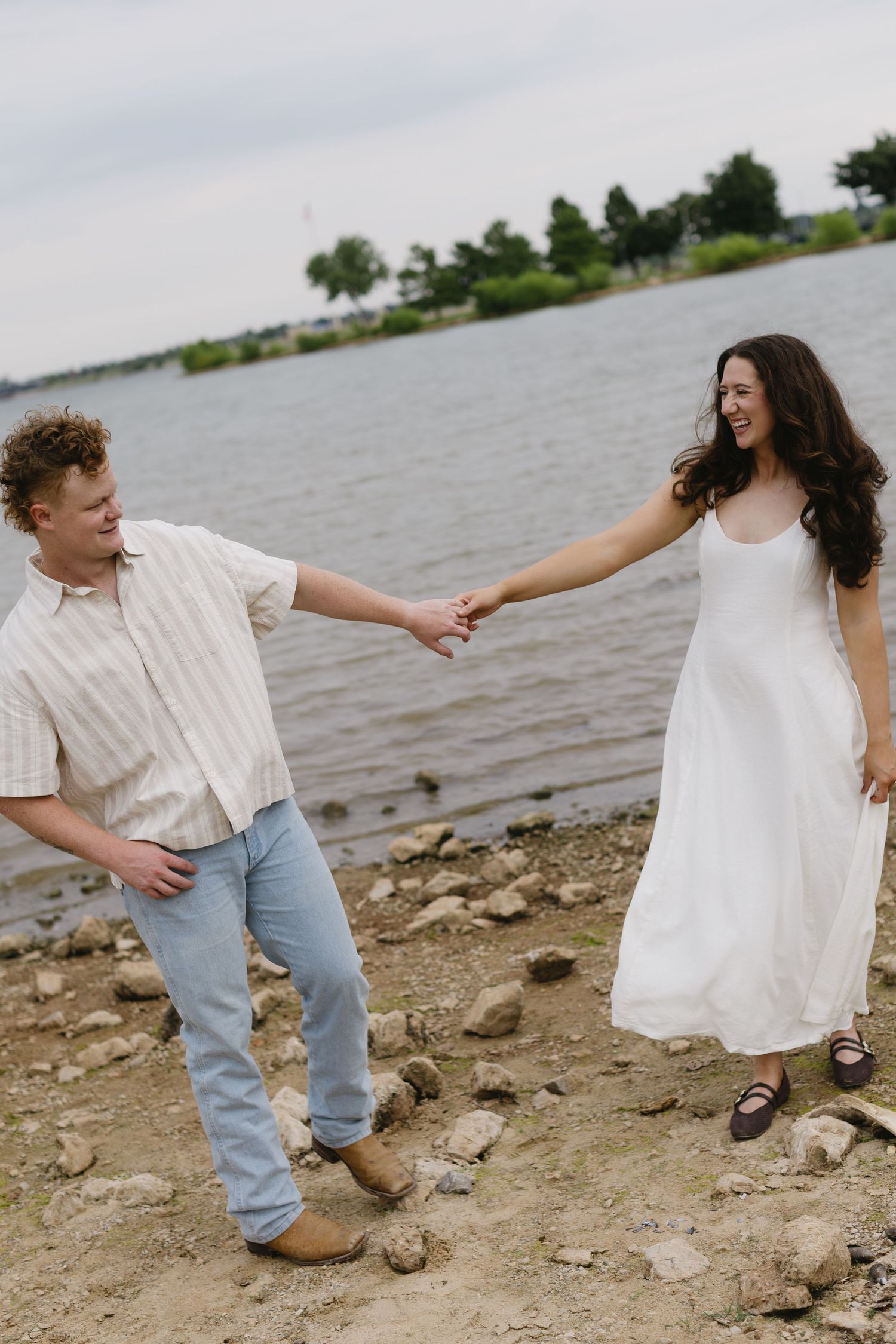 A playful sequence of a couple dancing and holding hands on a rocky beach beside a lake.