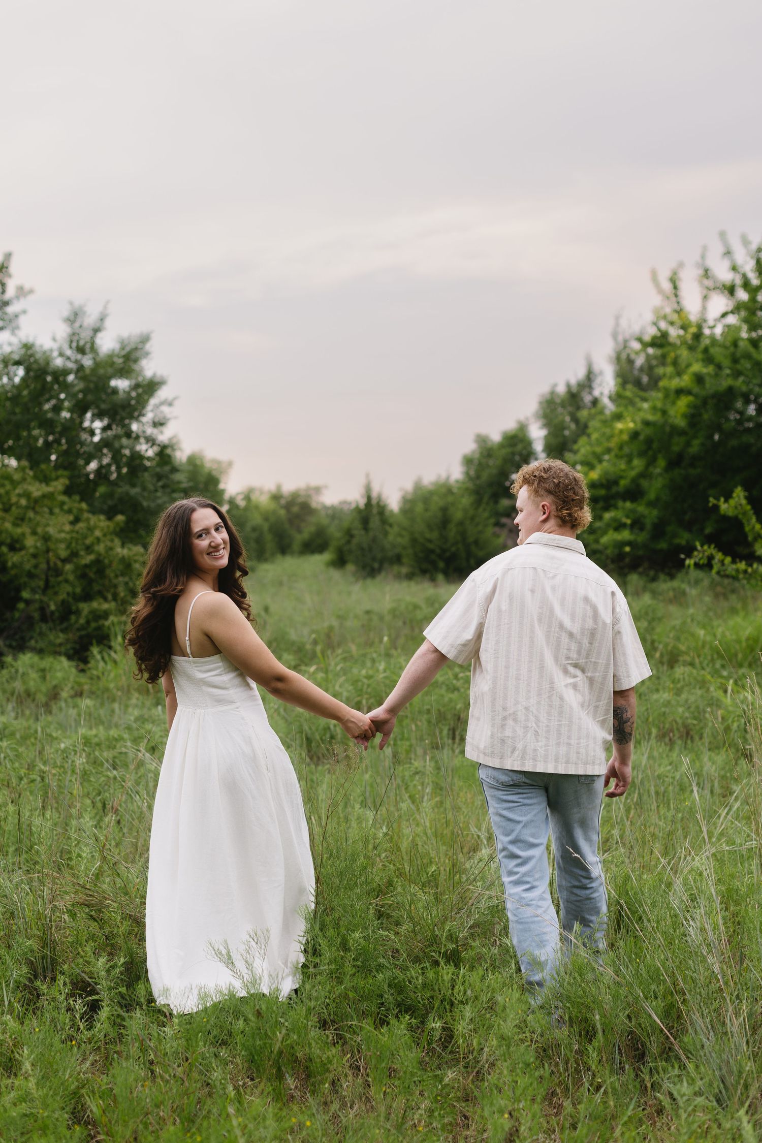 A couple in white clothing walks hand-in-hand through a grassy meadow.
