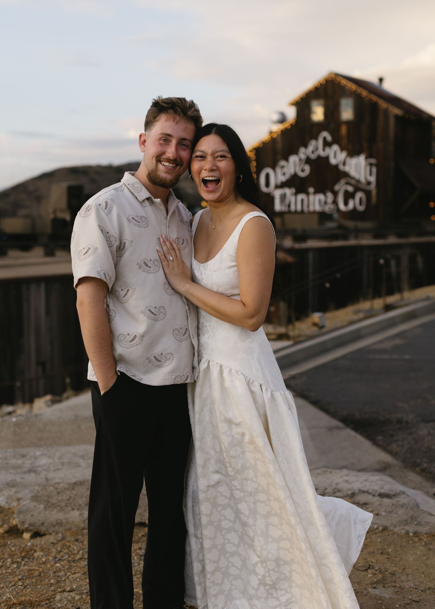 Couple in vintage-inspired outfits laughing together outside rustic buildings at sunset.