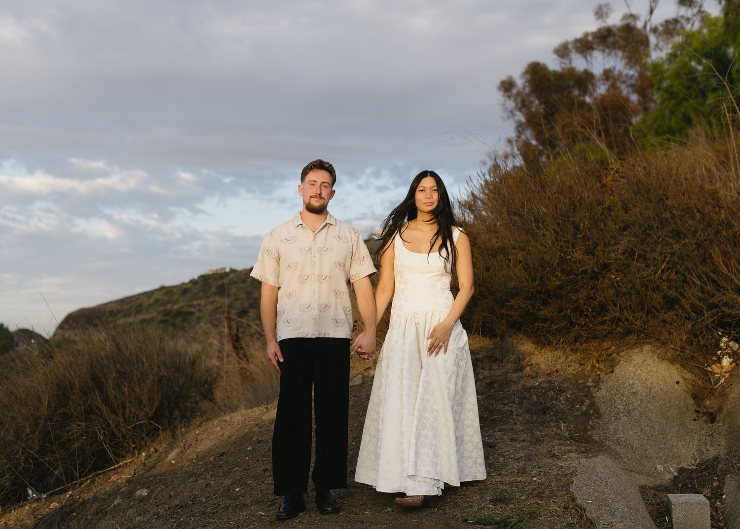 A couple in white outfits poses together on a scenic mountain trail at sunset.