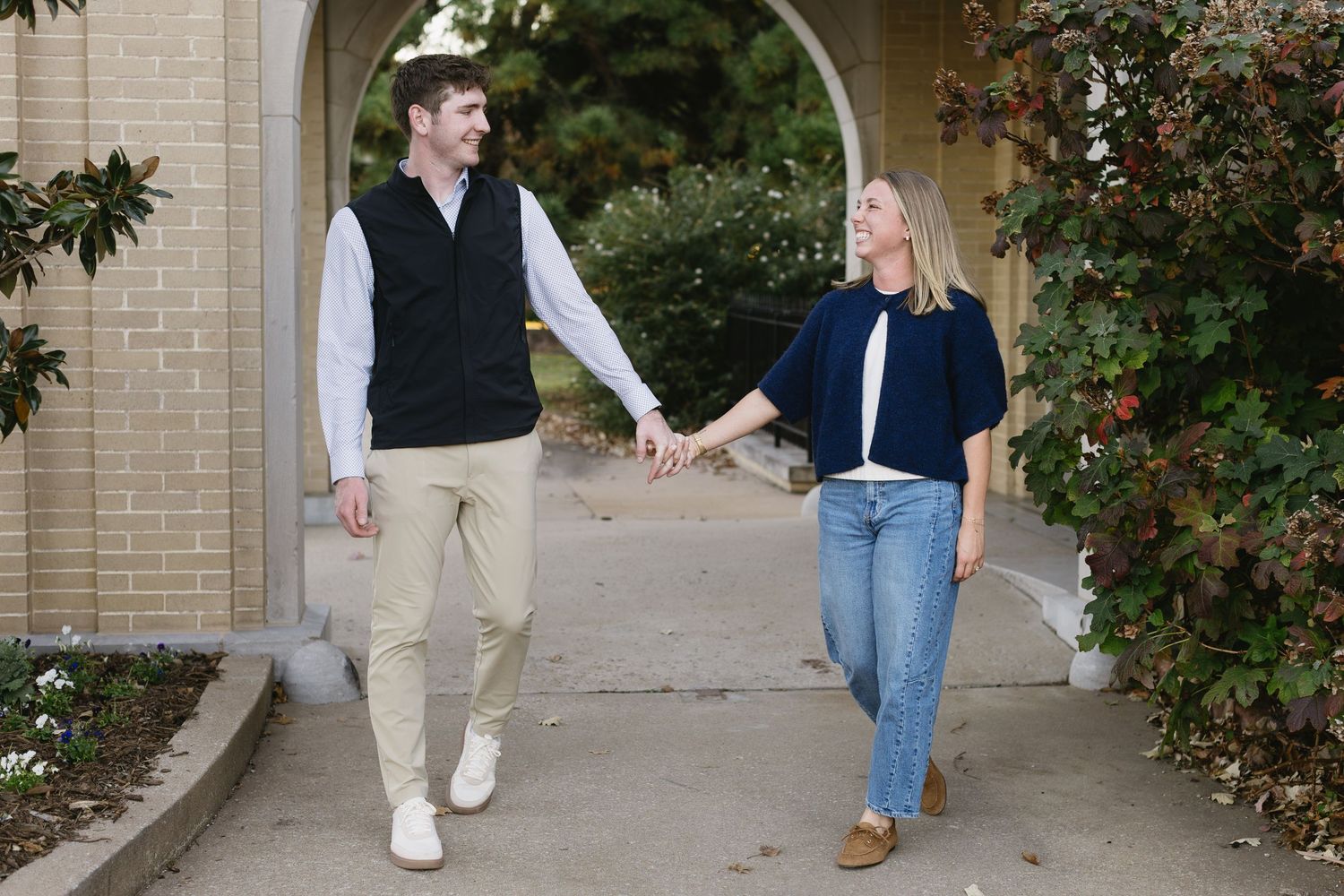 Two people walk together under an arched walkway near a brick building.