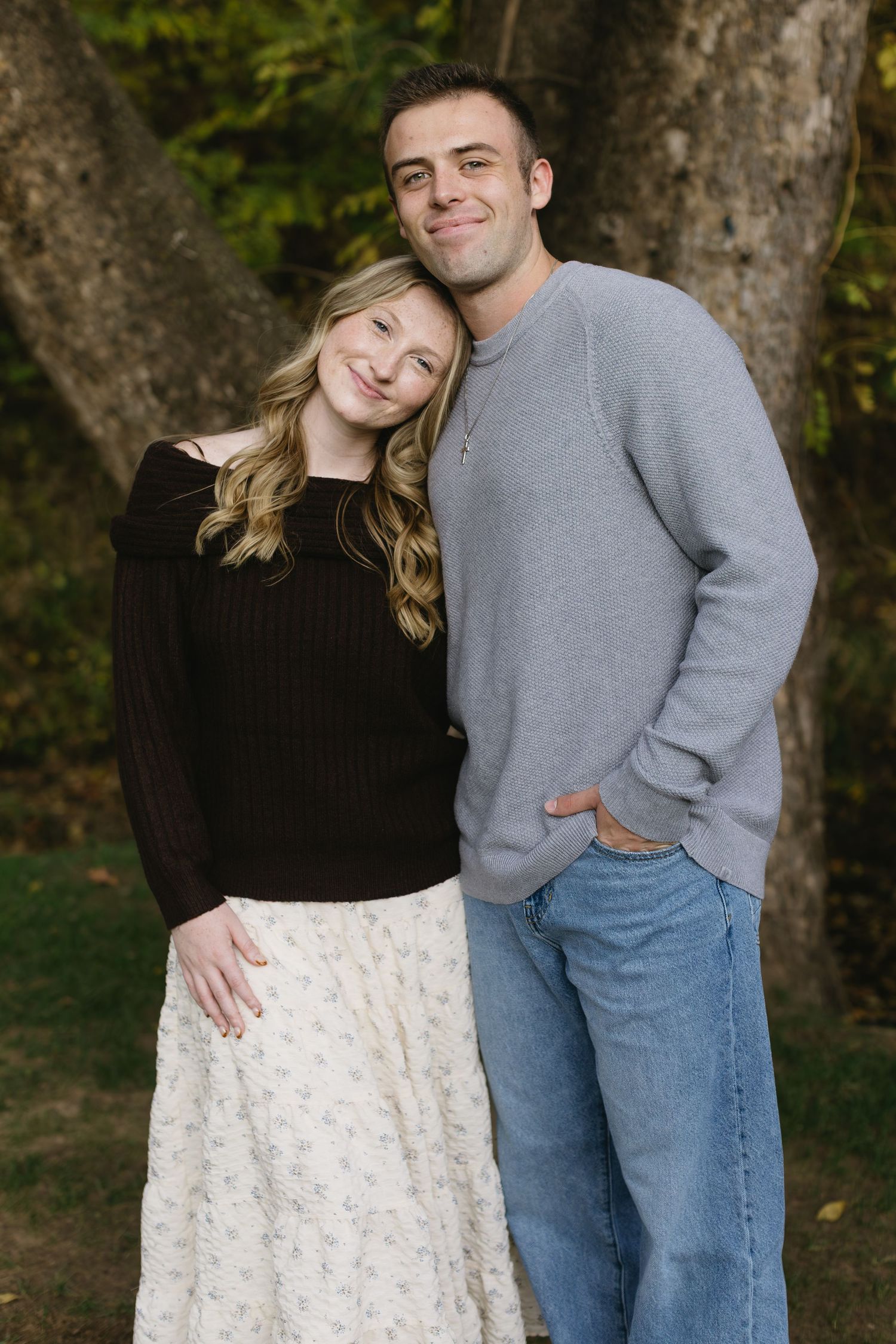 A couple poses together outdoors in casual attire with a grey sweater and floral skirt against a wooded backdrop.