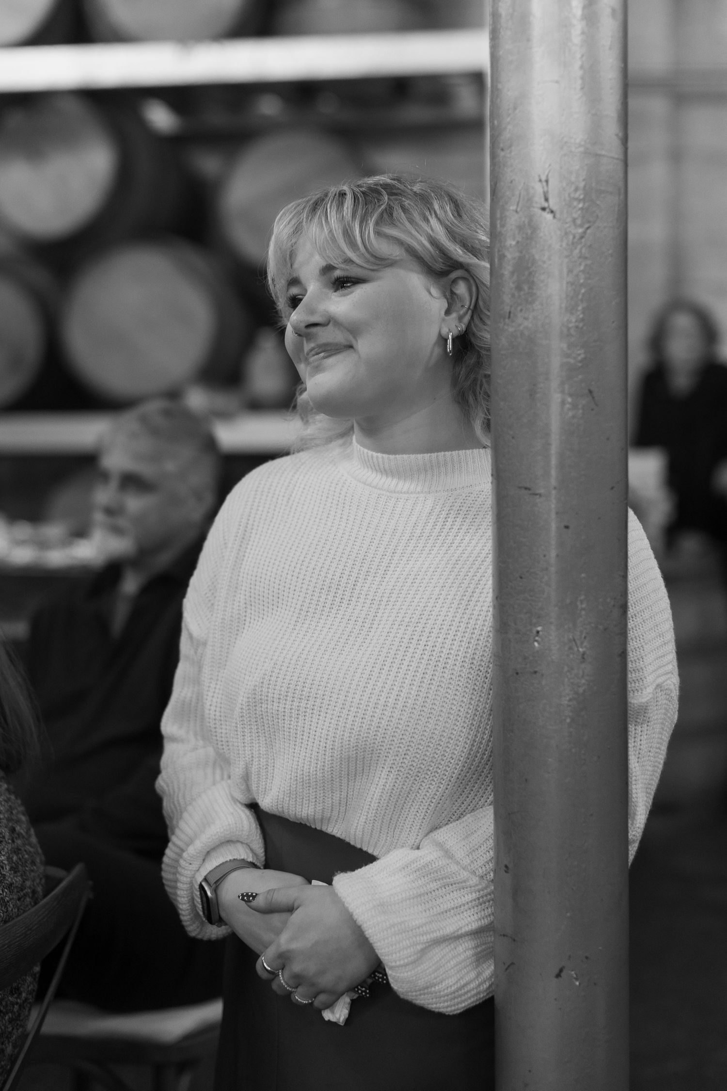 Person in white sweater smiling while leaning against a pole in monochrome photography.