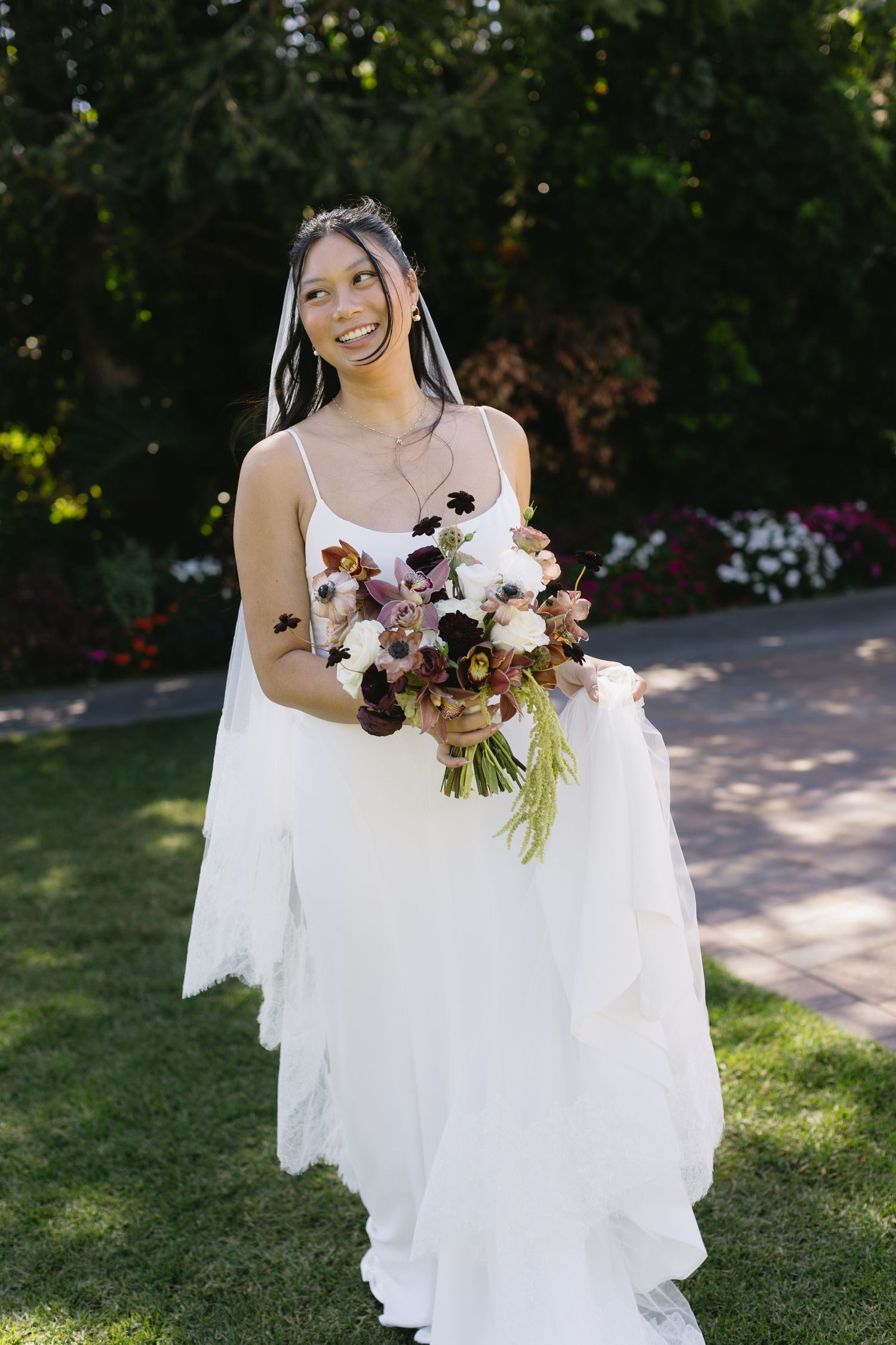 Joyful bride in flowing white gown holds burgundy and white bouquet in garden setting.