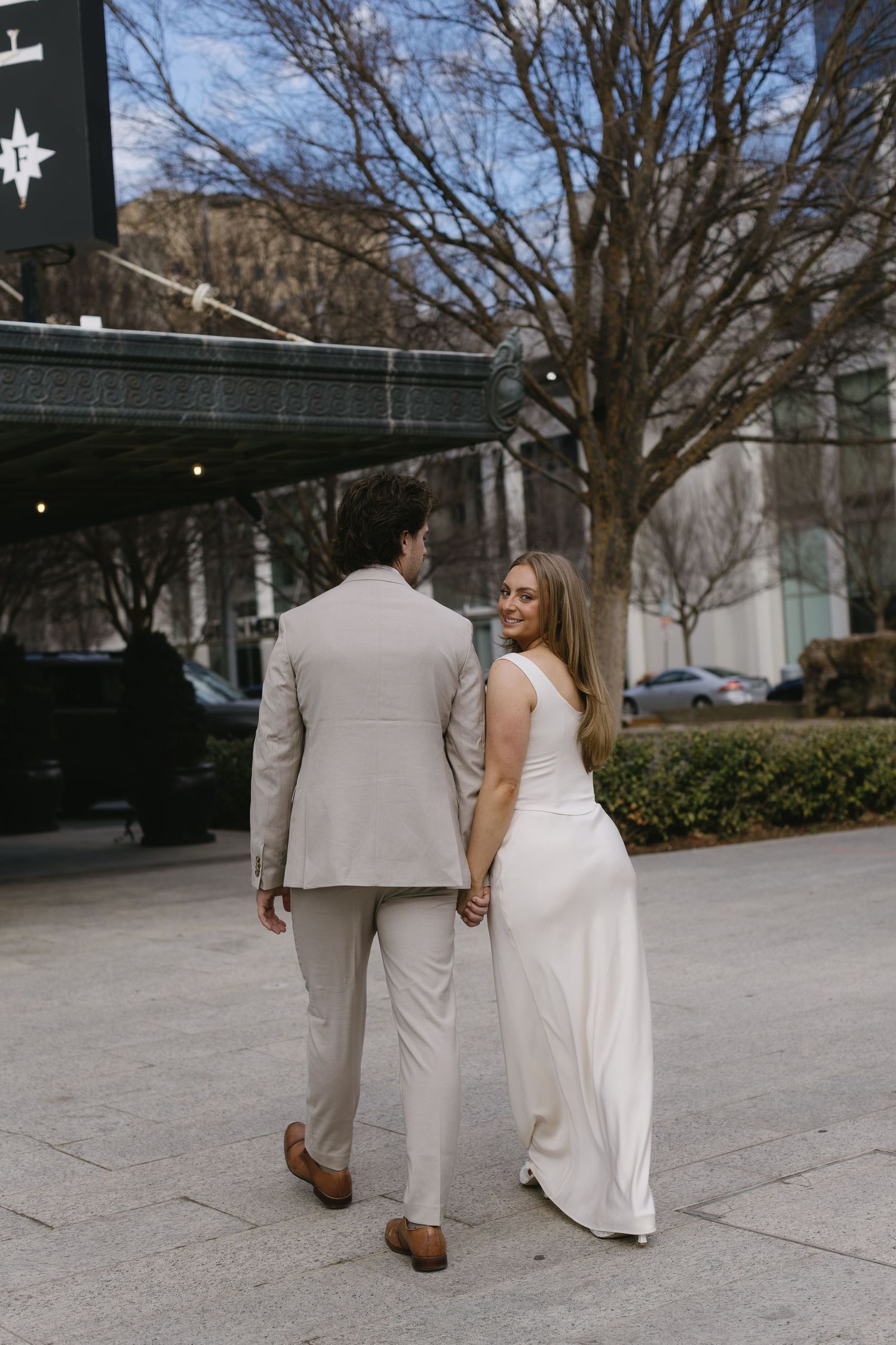 A couple walks together outdoors wearing white and beige formal attire.