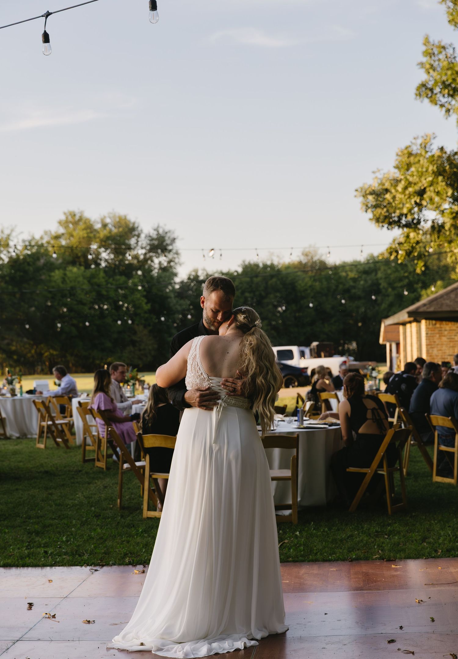Wedding reception outdoors at sunset with string lights, showing a couple dancing on a wooden dance floor.