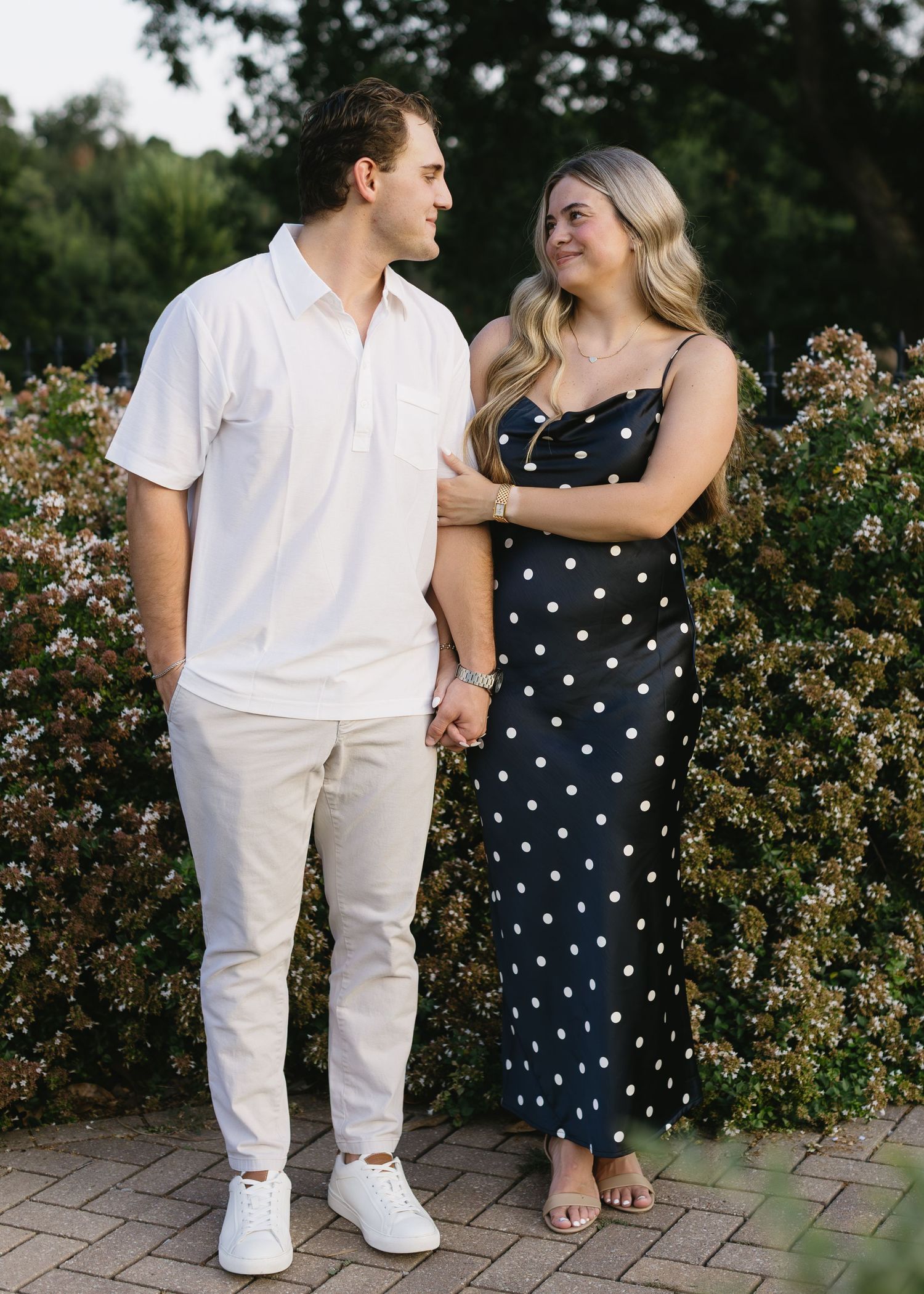 A couple in contrasting polka dot dress and white outfit poses near garden shrubs.