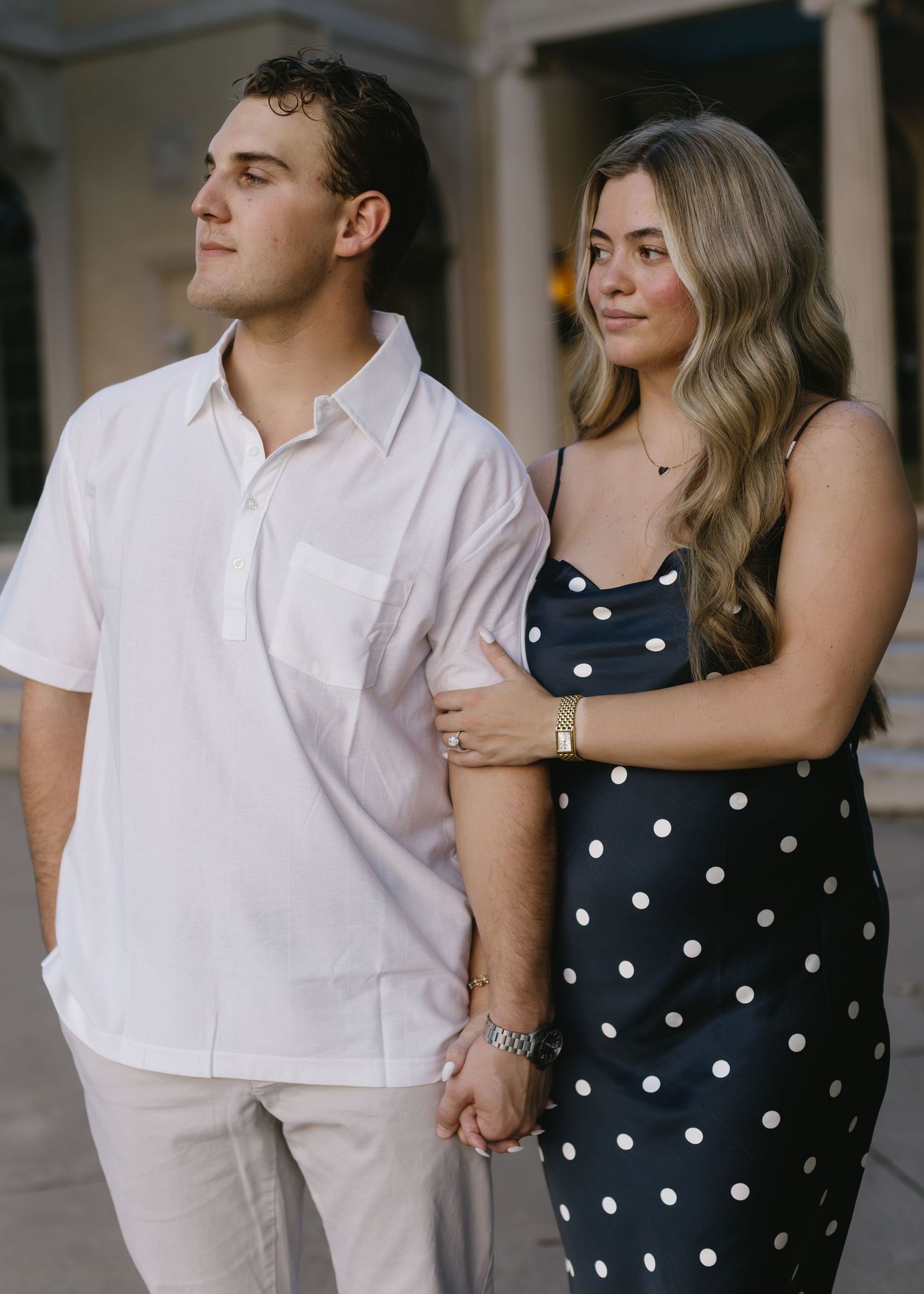 Couple in coordinated outfits share tender moment against classical architecture backdrop.