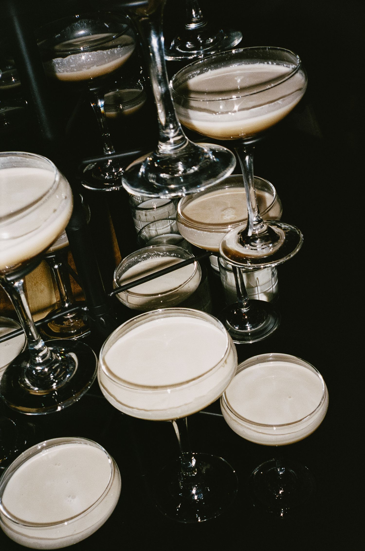 Multiple champagne coupes arranged on dark surface with frothy drinks.
