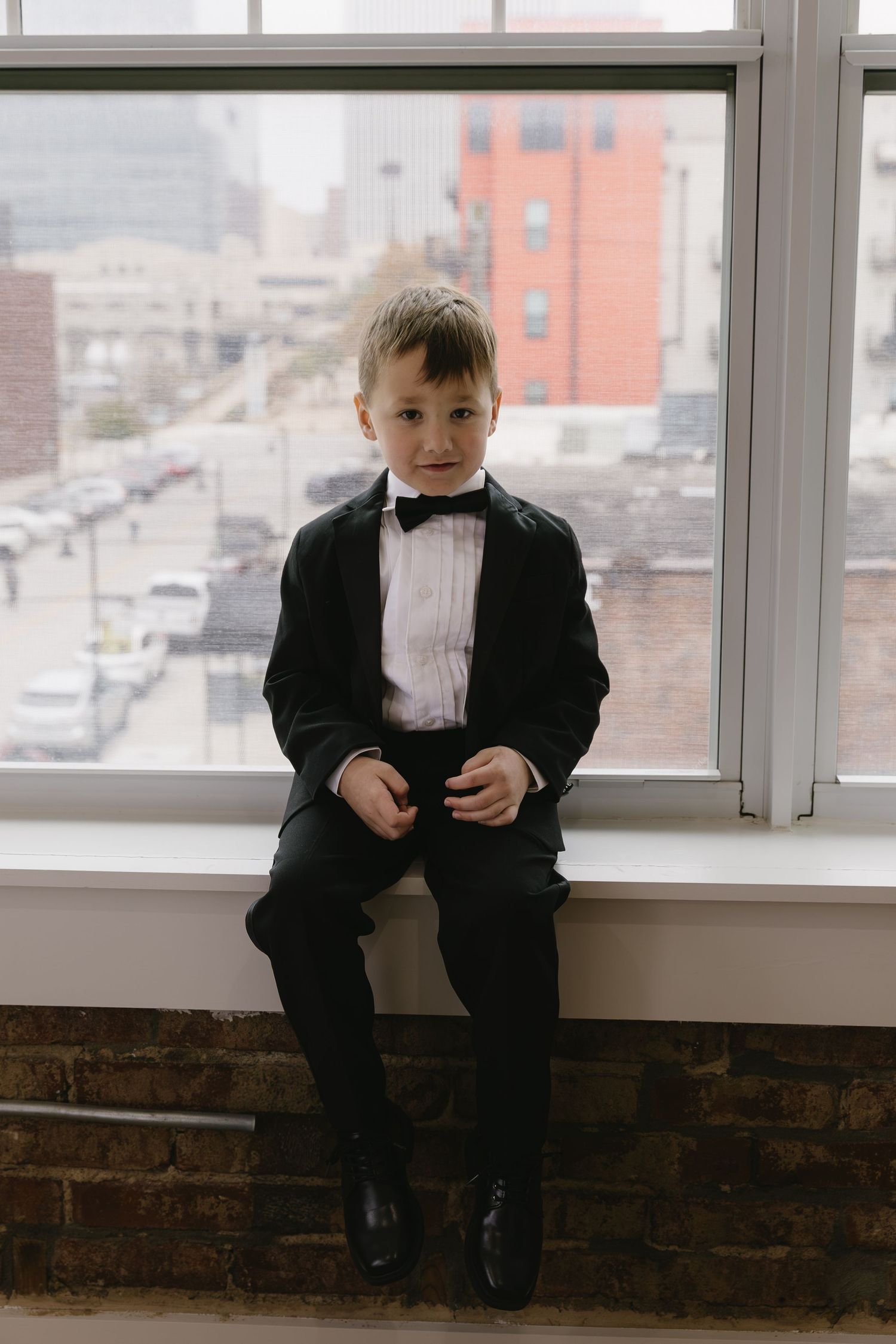 Young ring bearer in a black tuxedo sits alone on a windowsill looking at a phone.