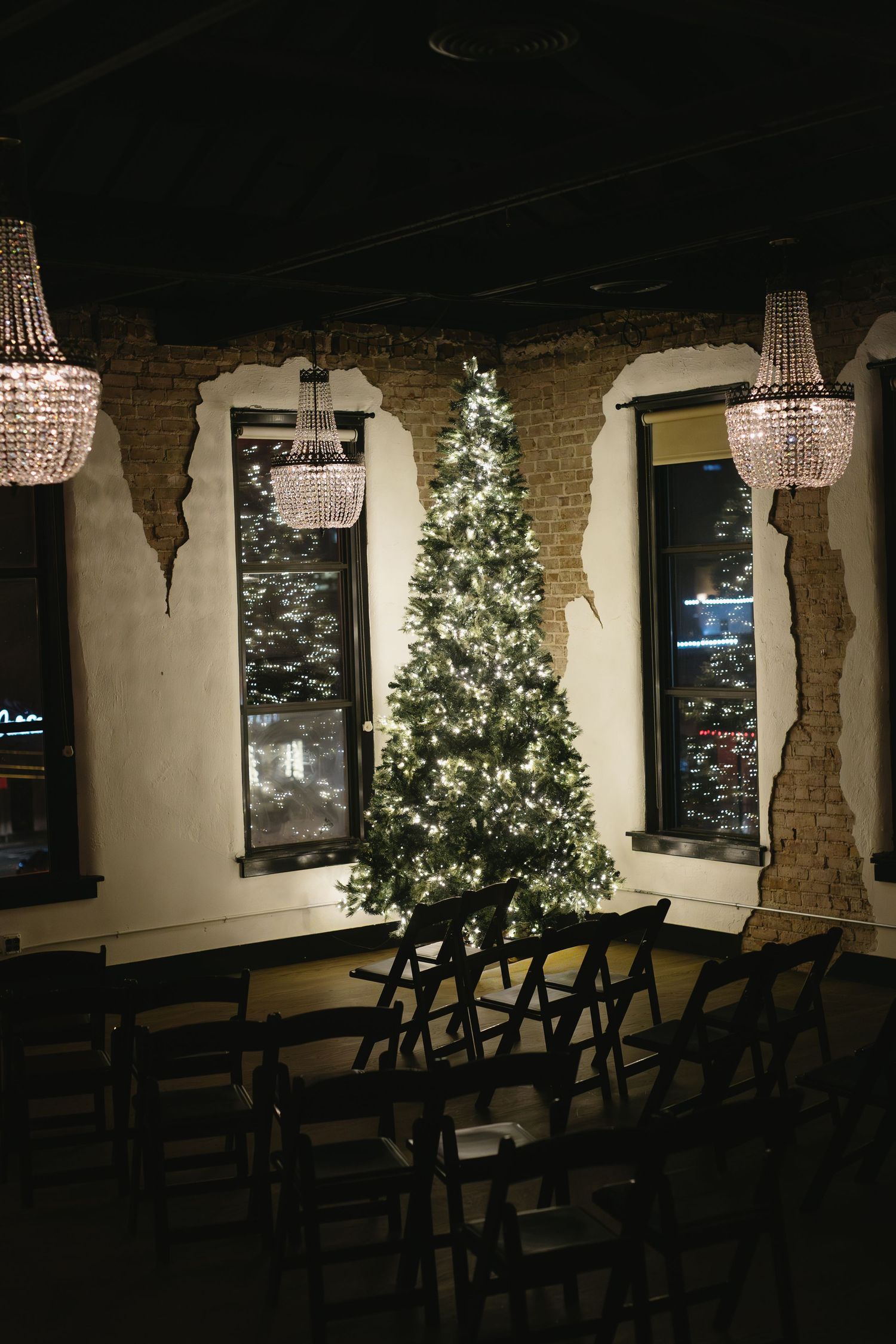 Illuminated Christmas tree stands between crystal chandeliers in a rustic venue with exposed brick walls.