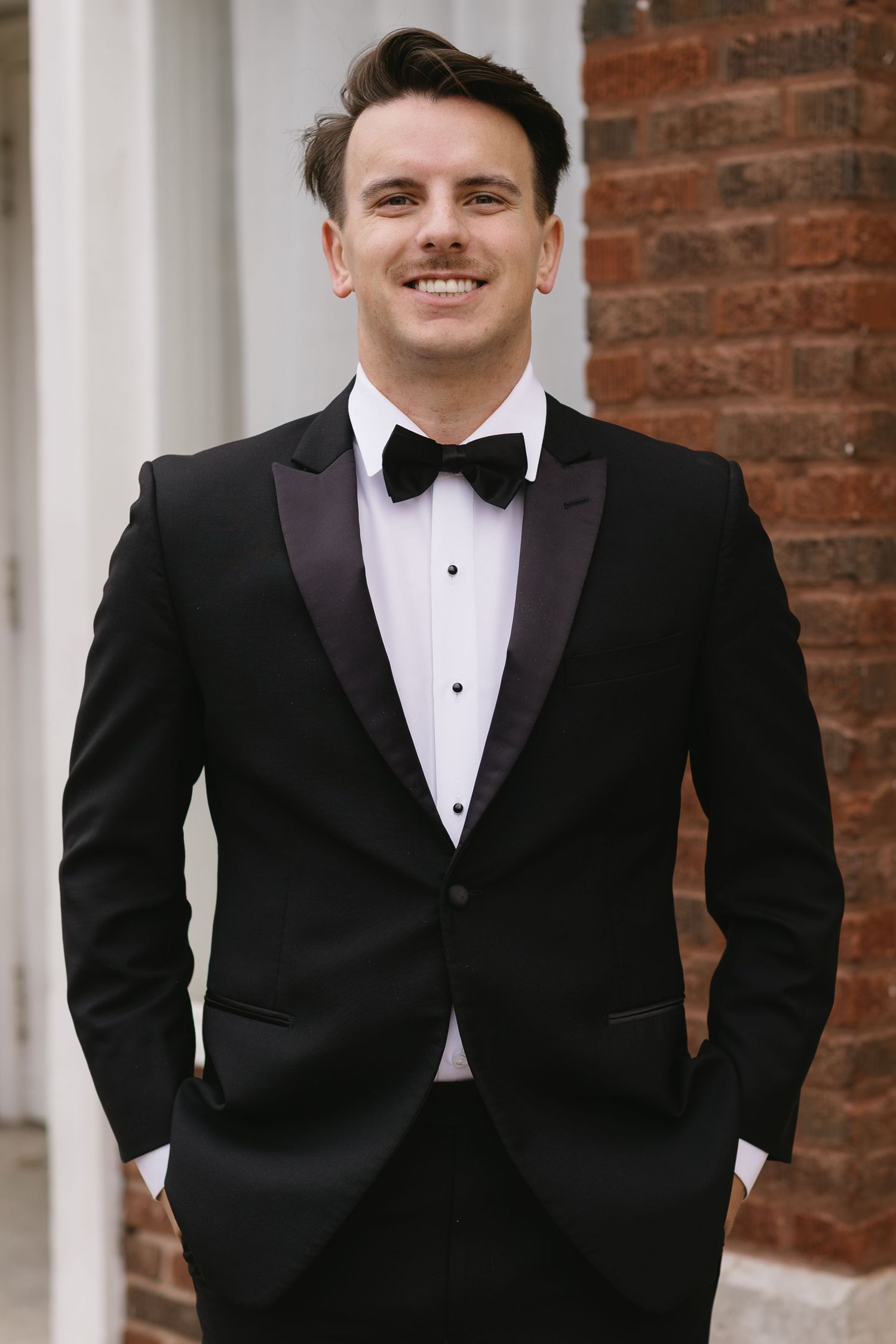 A groom in a classic black tuxedo and bow tie poses against a brick wall.