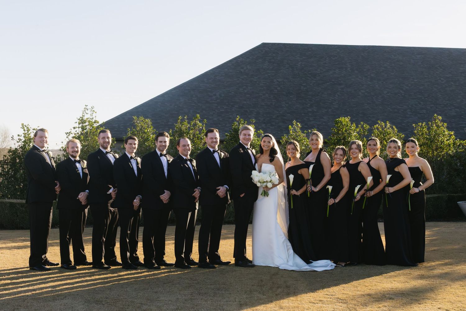 Large wedding party in black and white formal attire poses outdoors for group photo.