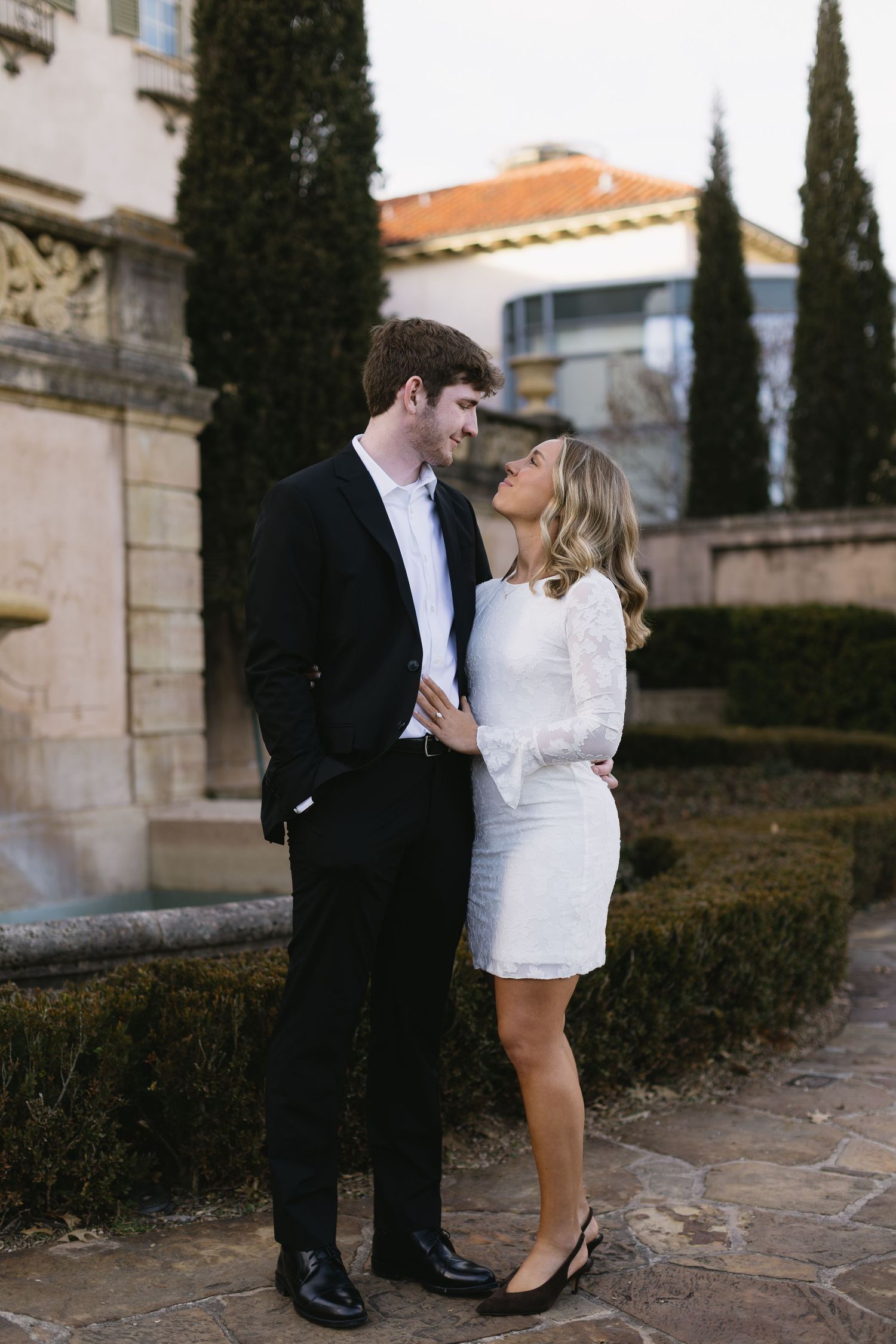 A romantic couple shares an intimate moment against a stone wall backdrop with cypress trees.