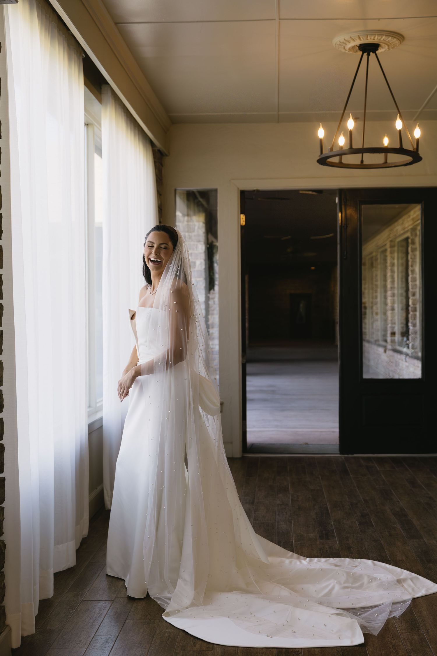 Bride in flowing white gown and veil poses by window in rustic indoor setting.