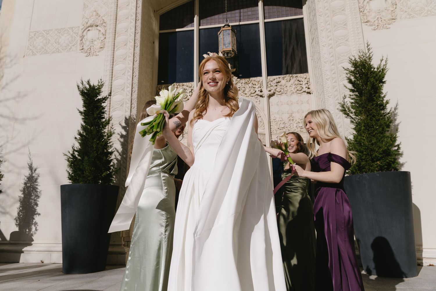Wedding party walking outside elegant building with bride in white gown and bridesmaids in purple.