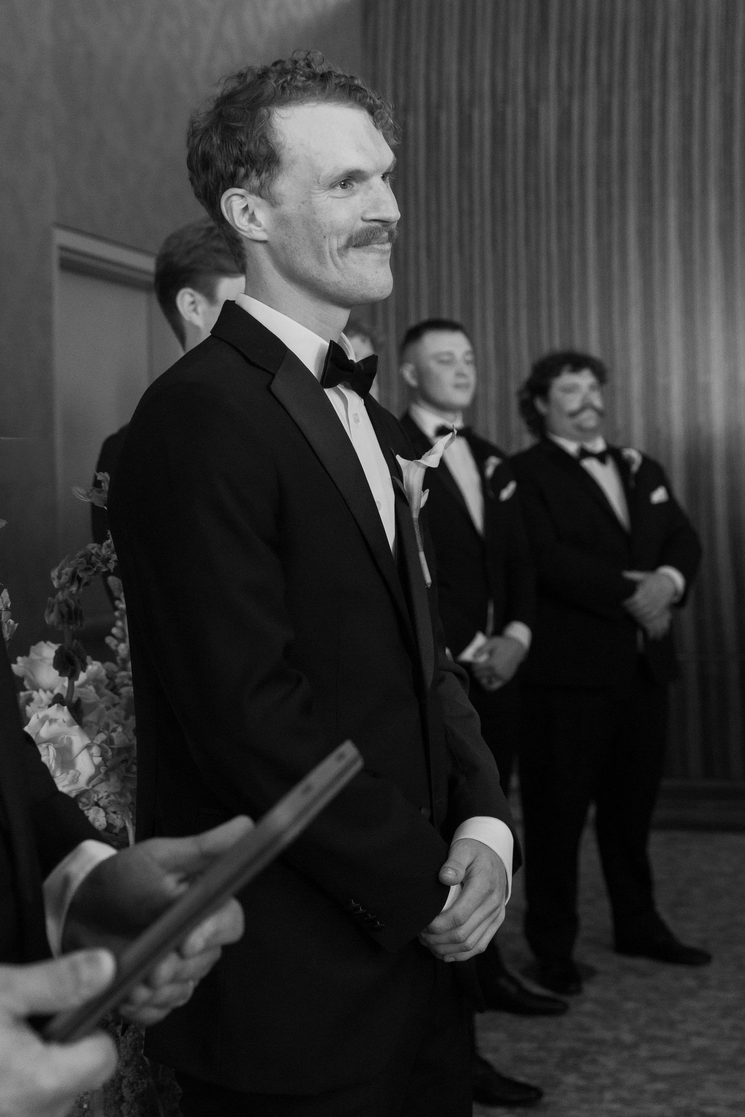 Guests in formal black tie attire mingle and laugh during an elegant indoor reception.