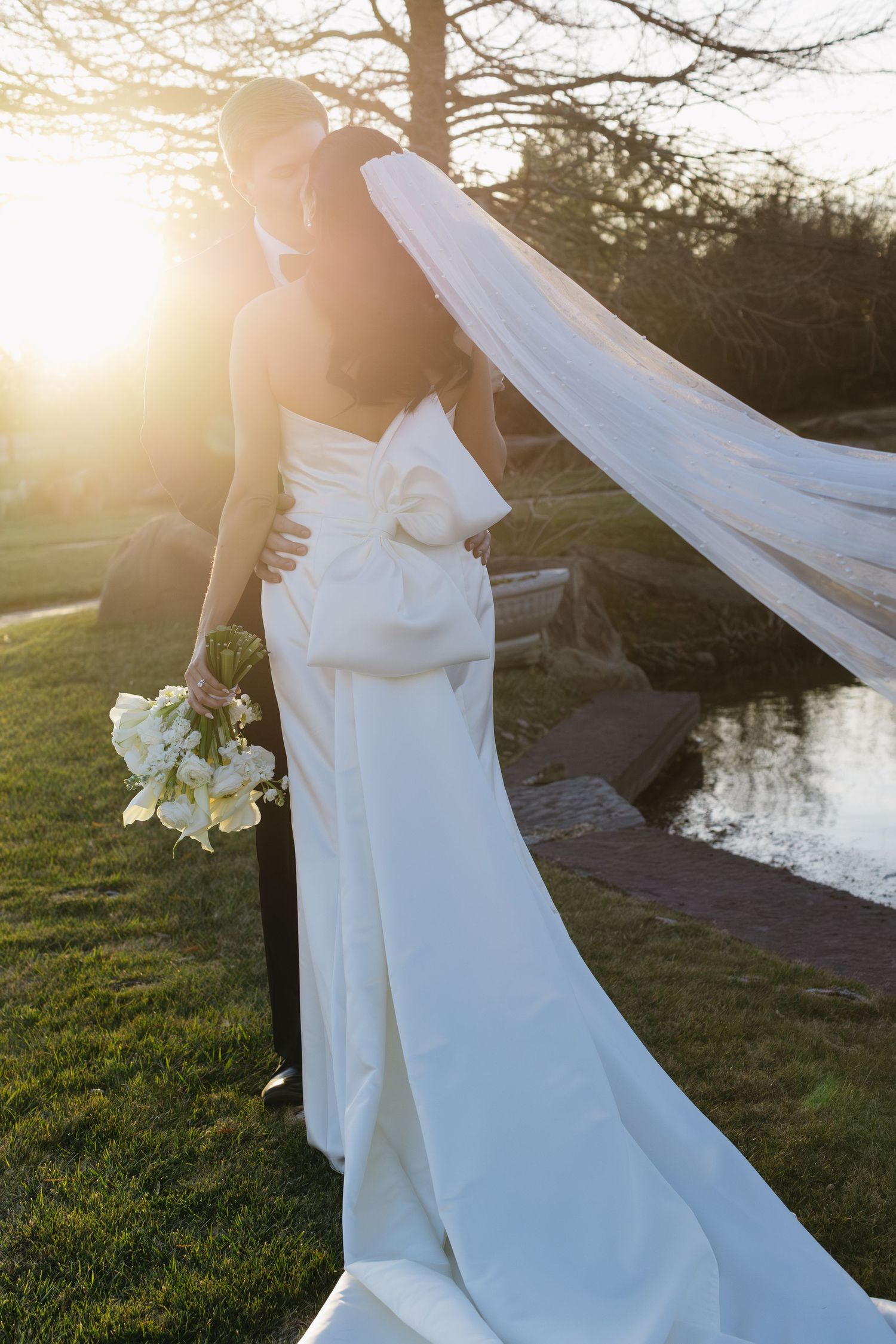 A flowing white wedding dress catches sunlight by a pond during golden hour.
