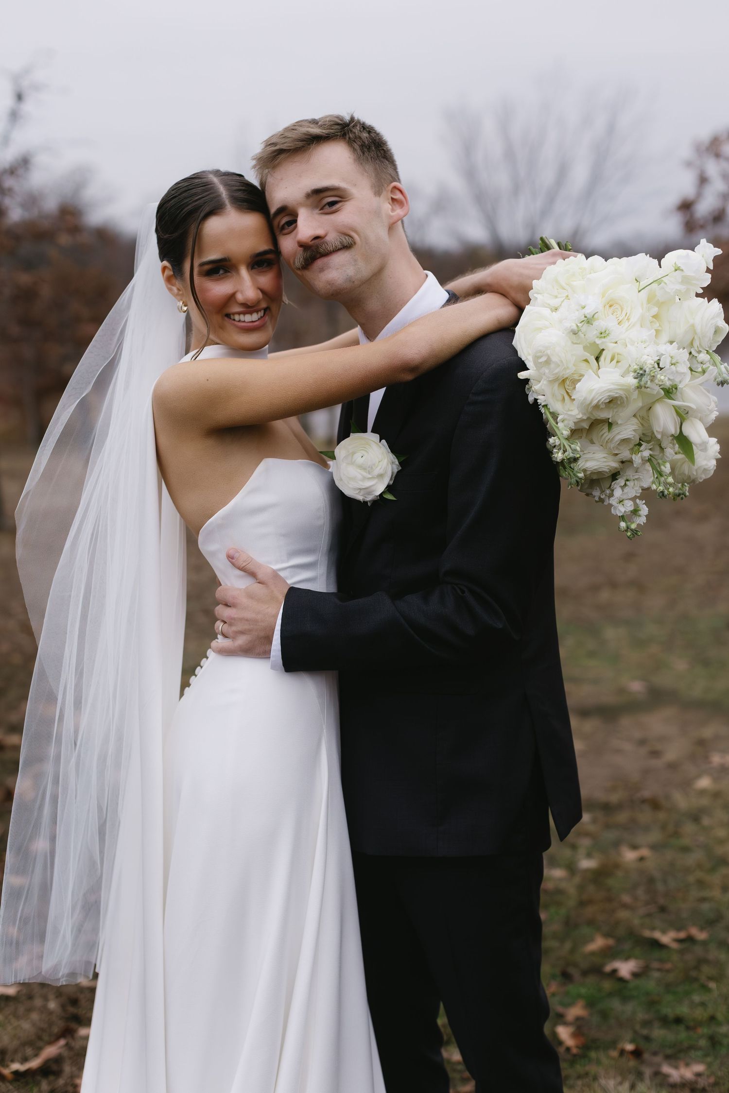 Couple embracing outdoors in formal wedding attire with white bouquet and veil.