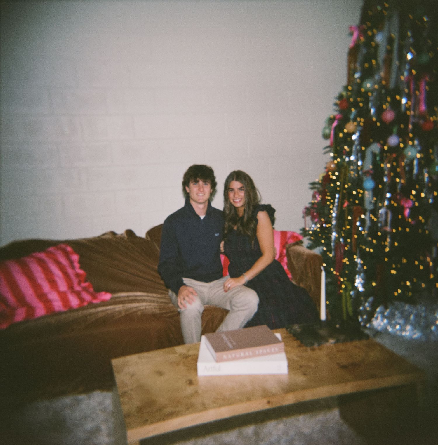People sit together on a couch in front of a decorated Christmas tree in a dimly lit living room.