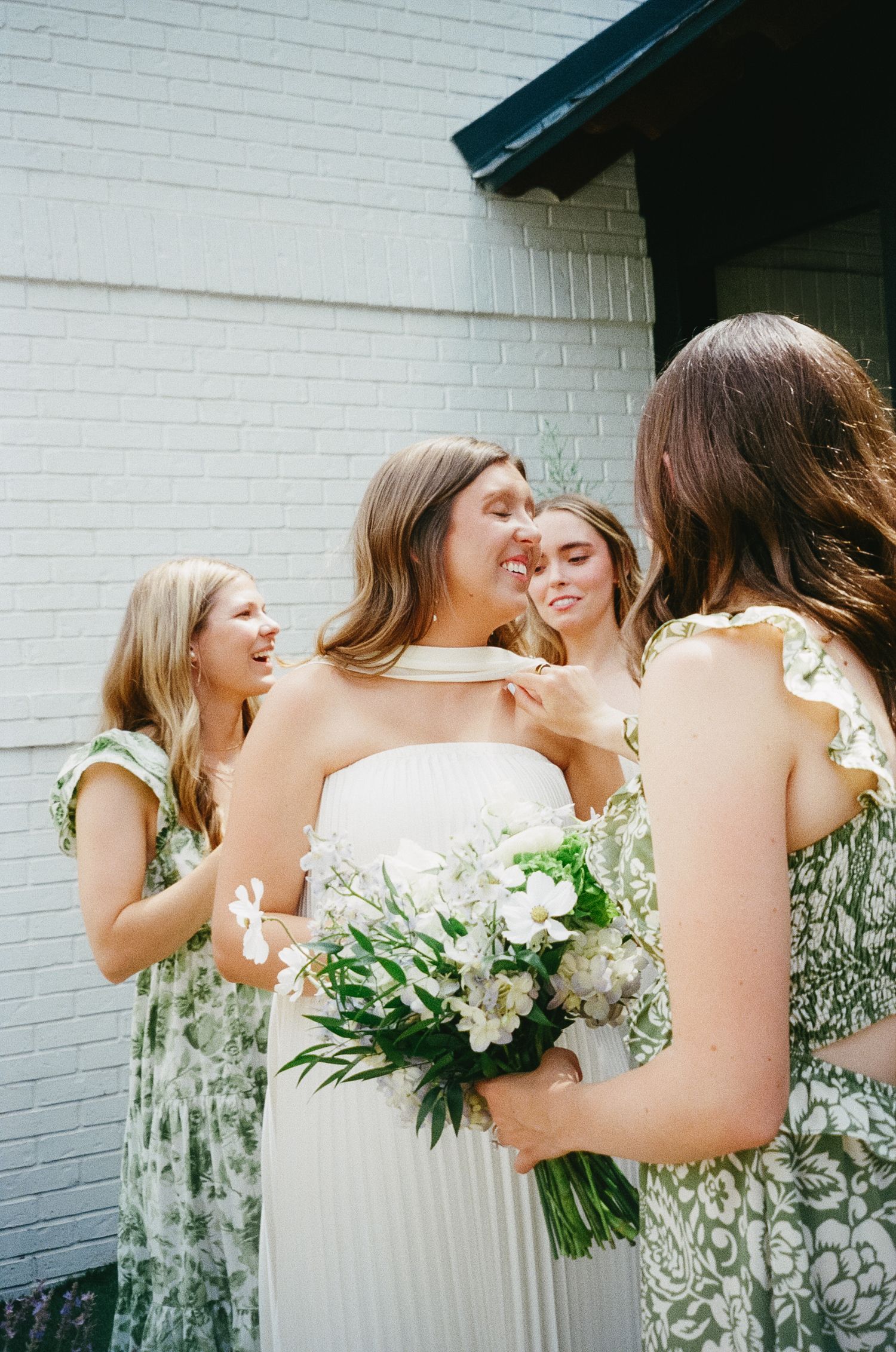 Bridesmaids in floral and navy dresses holding white bouquets against a white brick wall.