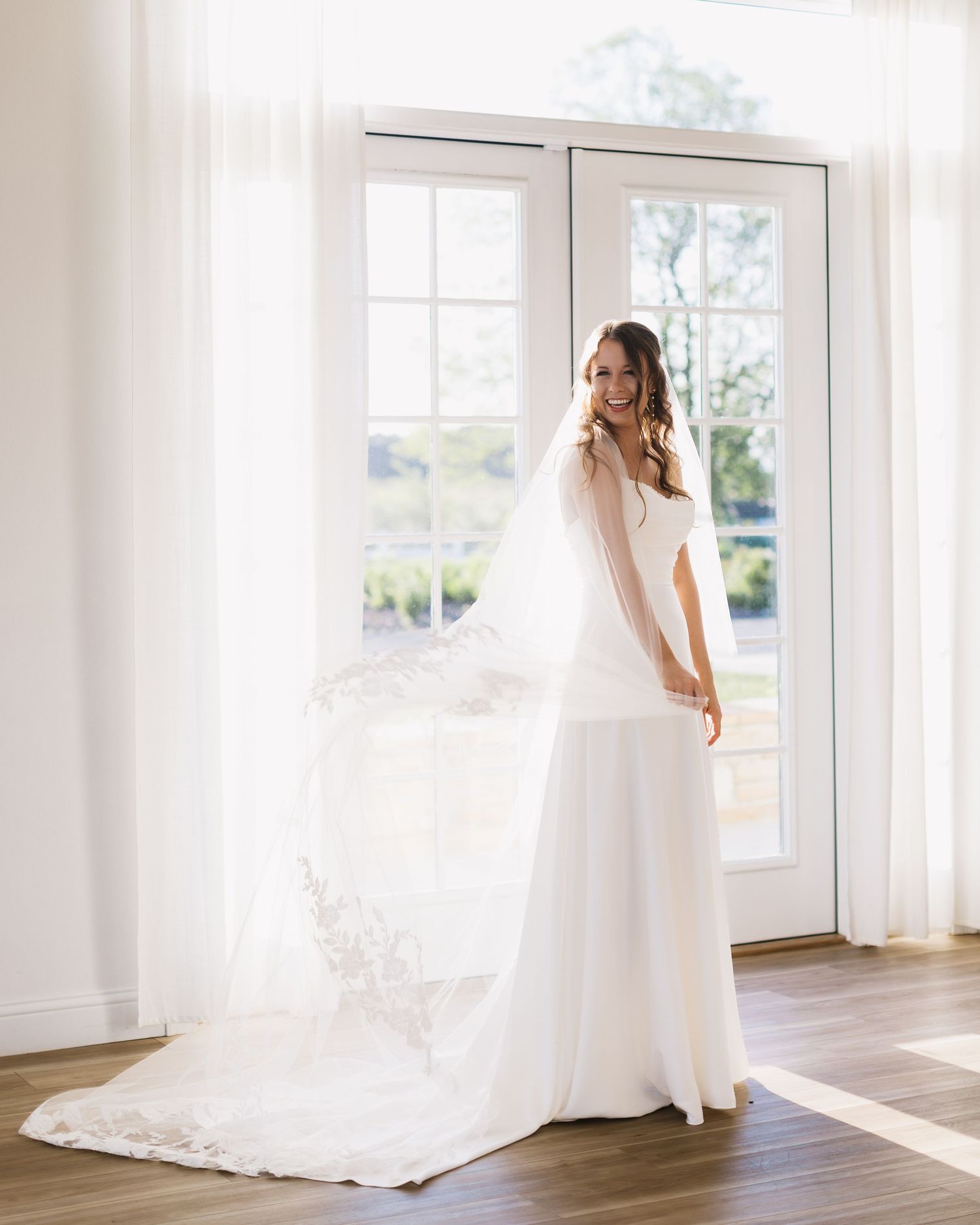 A bride in a flowing white wedding gown and veil stands in a sunlit room with bright white walls.