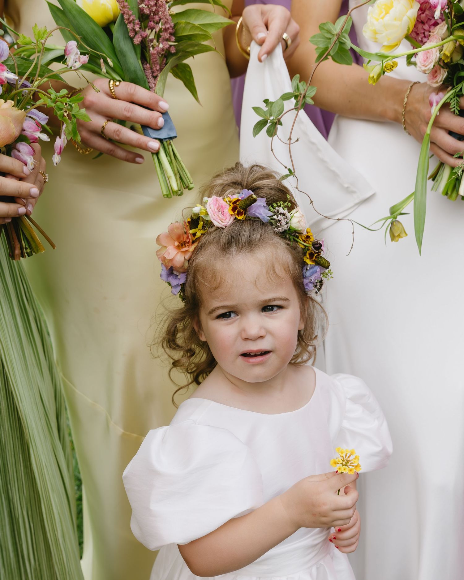 Young flower girl in white dress with floral crown smiles while holding bouquet among bridesmaids.