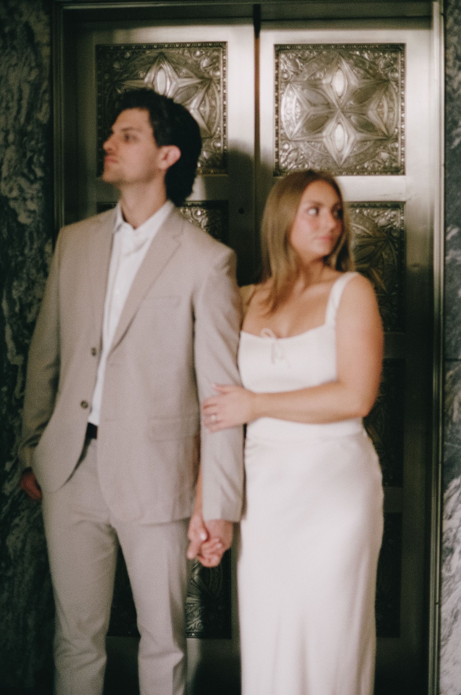 A couple in formal wear poses against a decorative wall with patterned details.