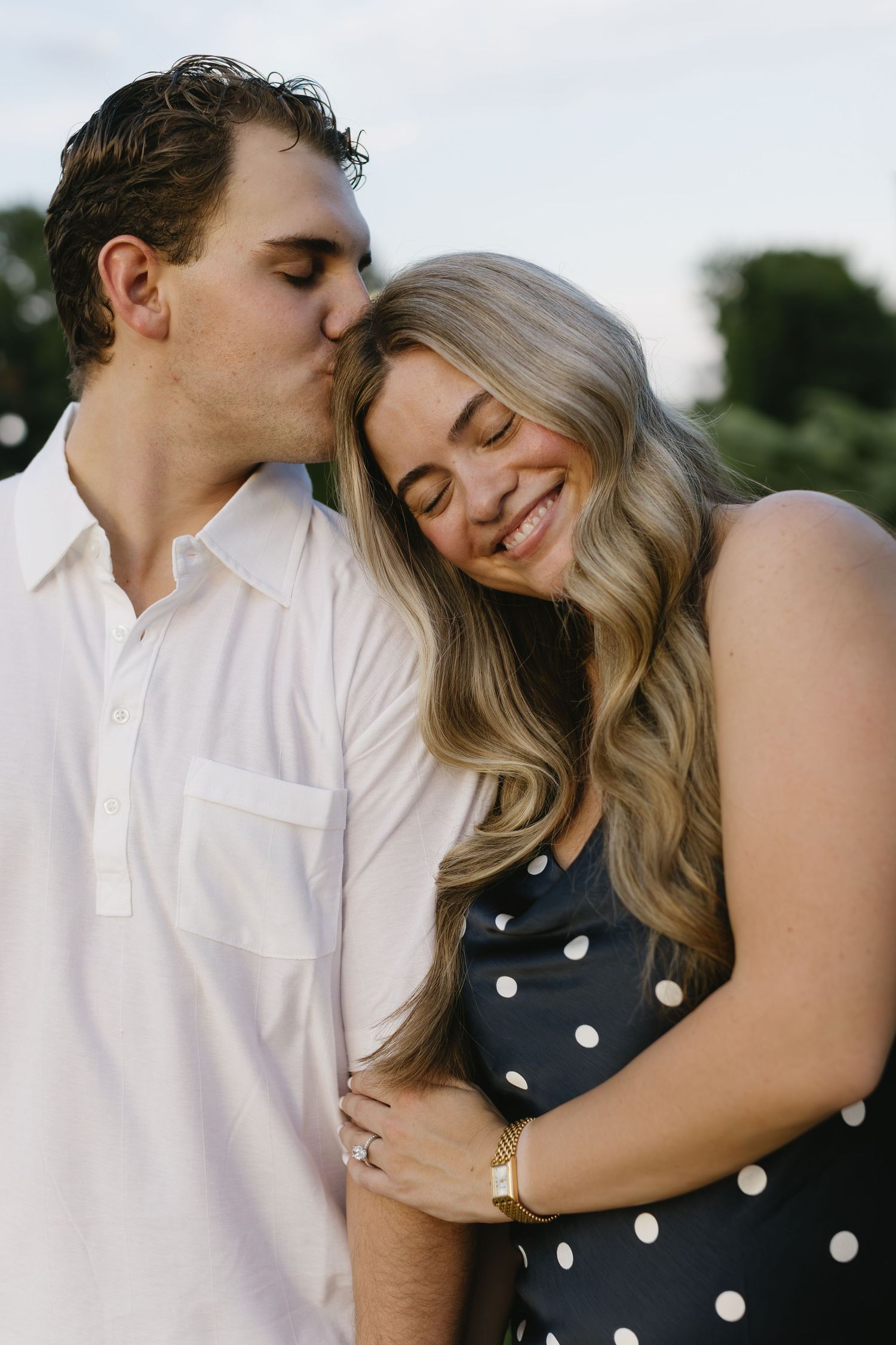 A romantic couple embraces outdoors, with her wearing a polka dot dress and him in a white shirt.