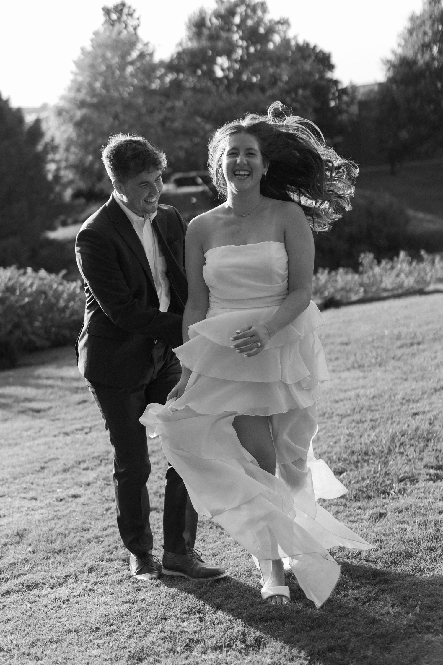 A joyful wedding couple running together on a grassy hillside in black and white photography.