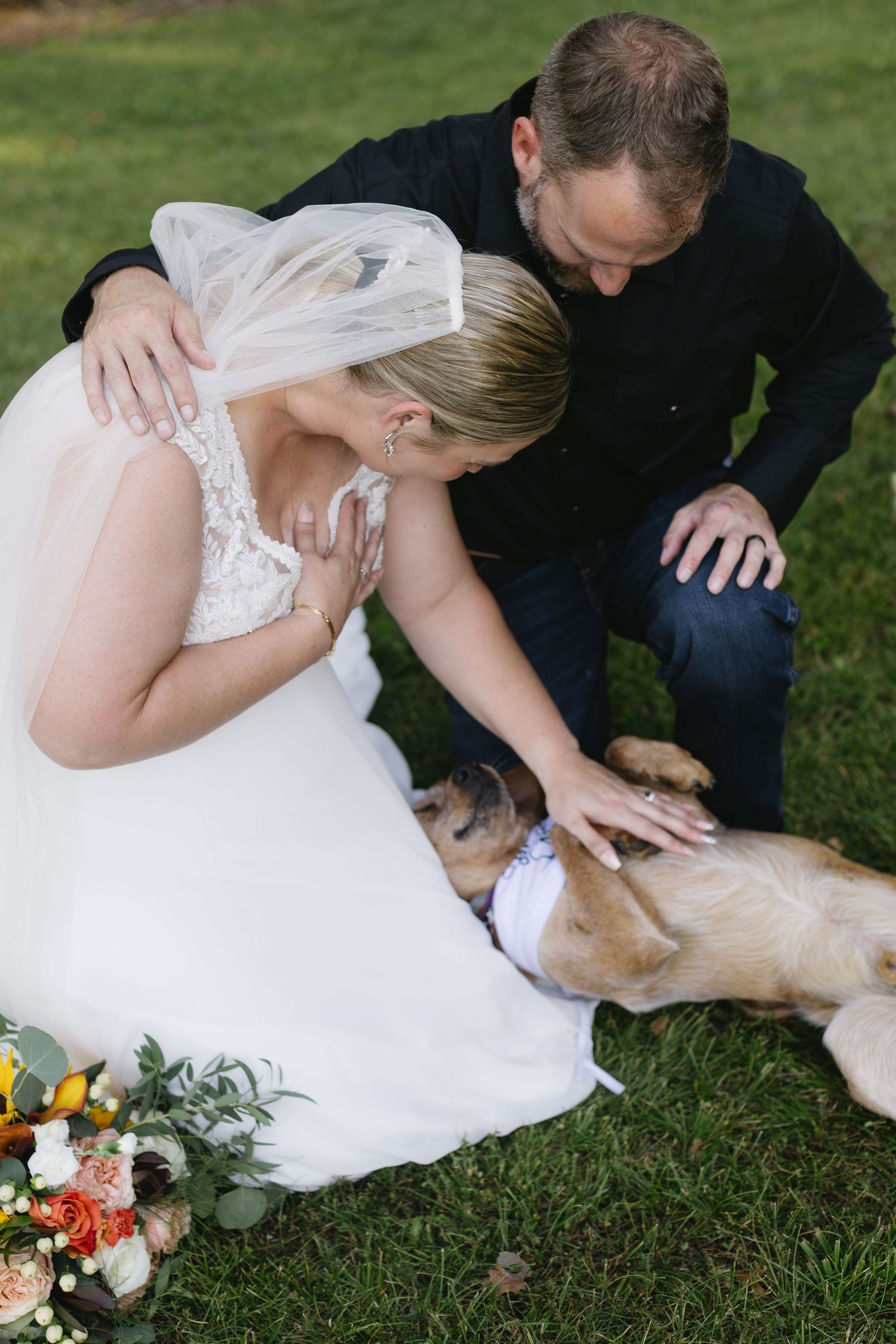 A bride in a white dress pets a dog on the grass while holding a floral bouquet.