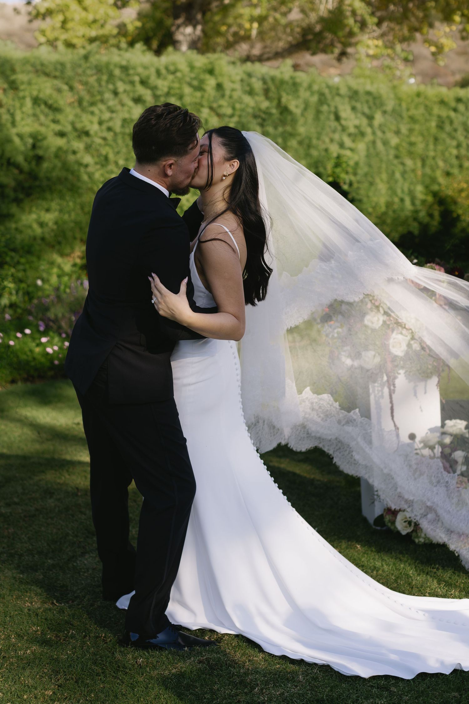 Romantic wedding couple kisses while bride's white veil flows dramatically in outdoor setting.