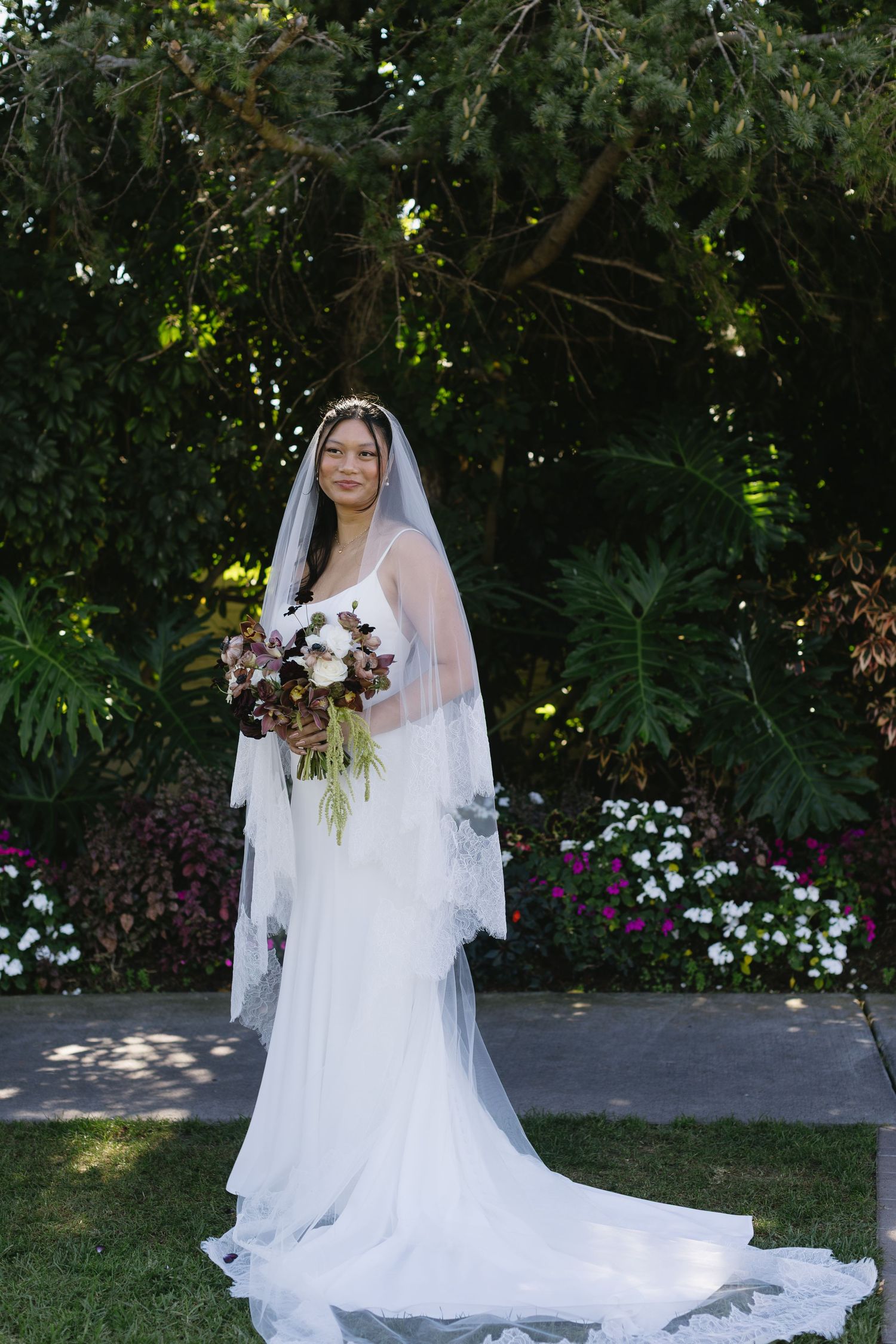Bride in elegant white gown and veil poses with bouquet against lush garden backdrop.