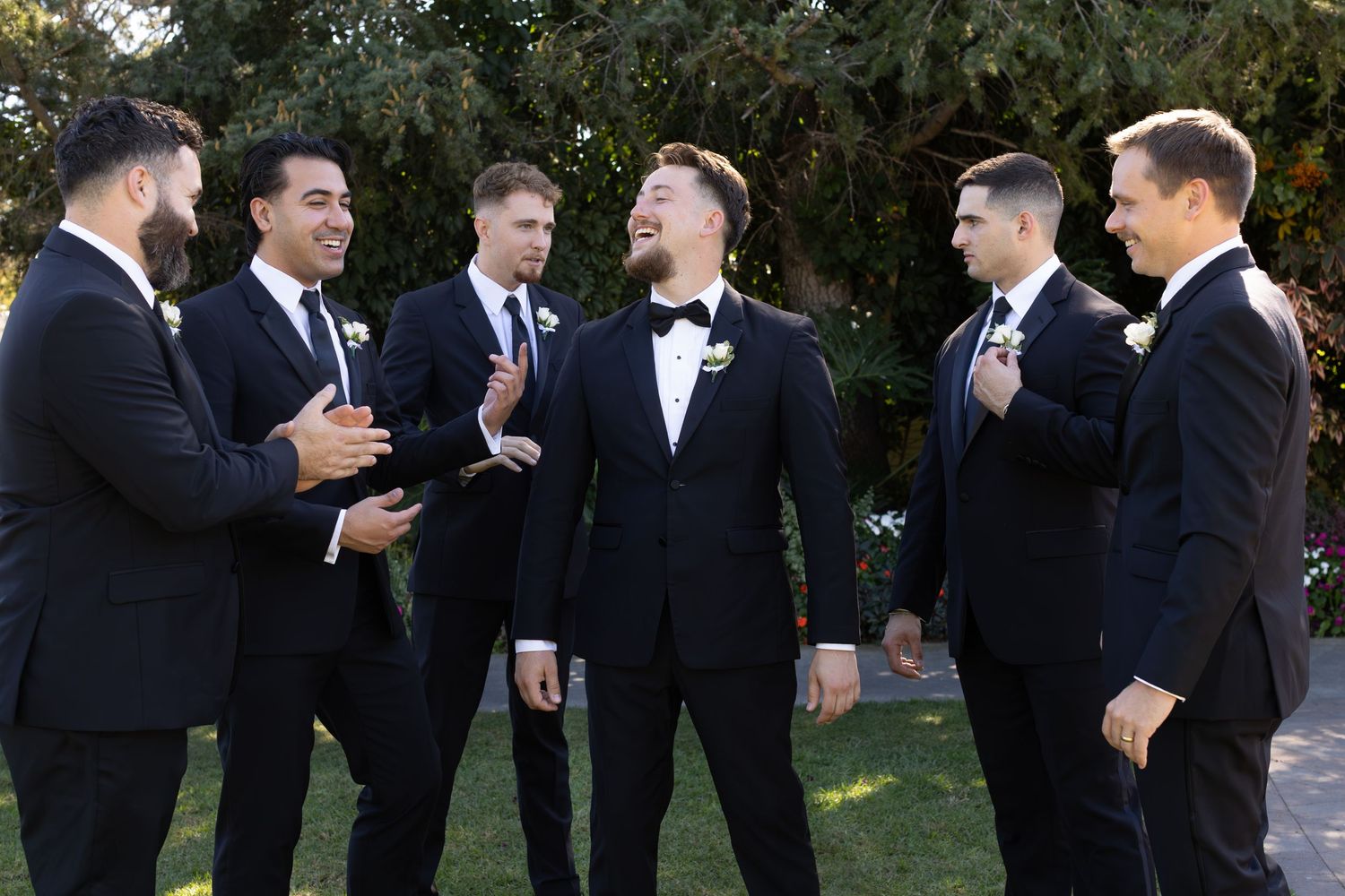 Groomsmen in black suits laughing together outdoors during wedding celebration.