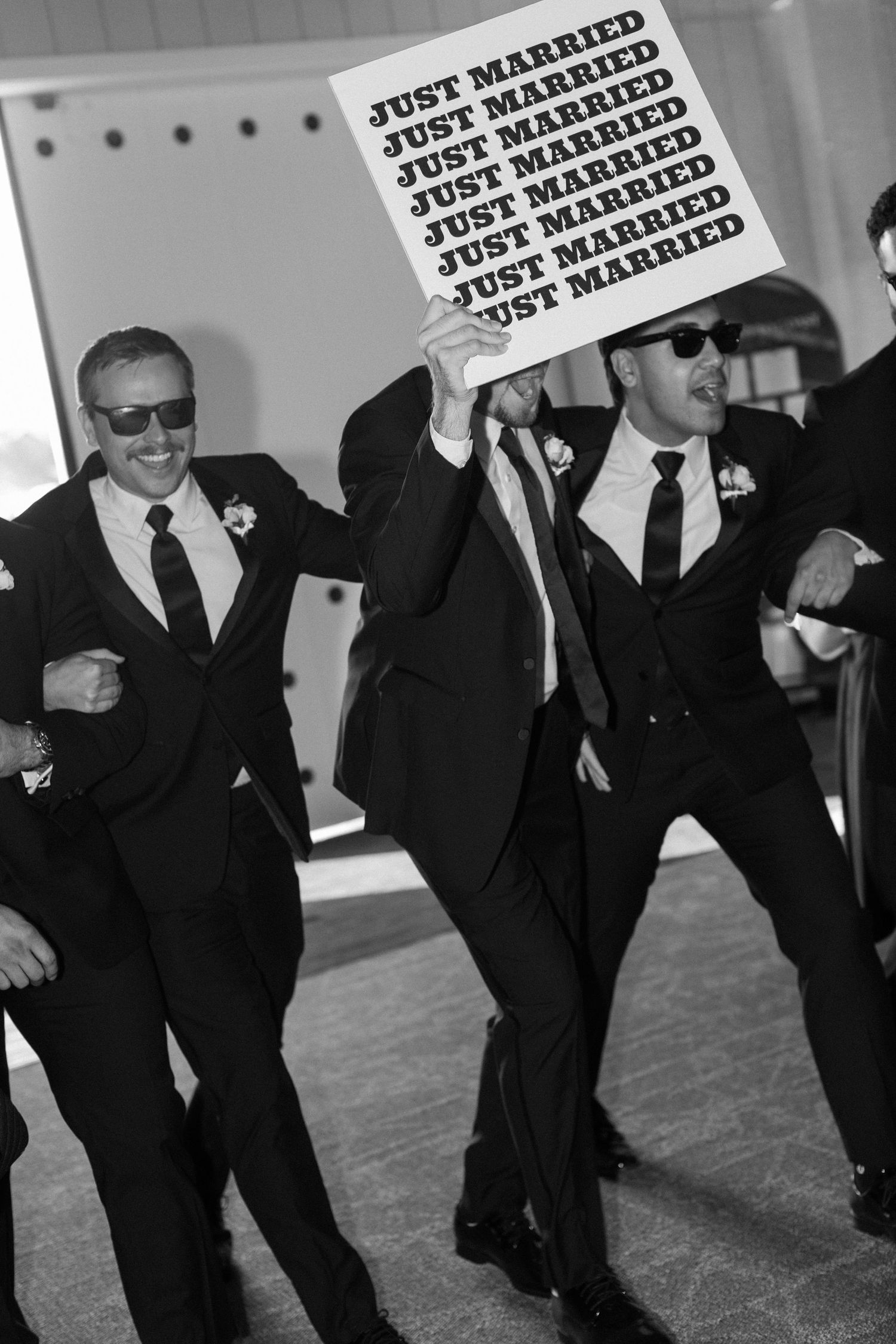 Wedding party holds 'Just Married' signs while dancing in black and white photo.
