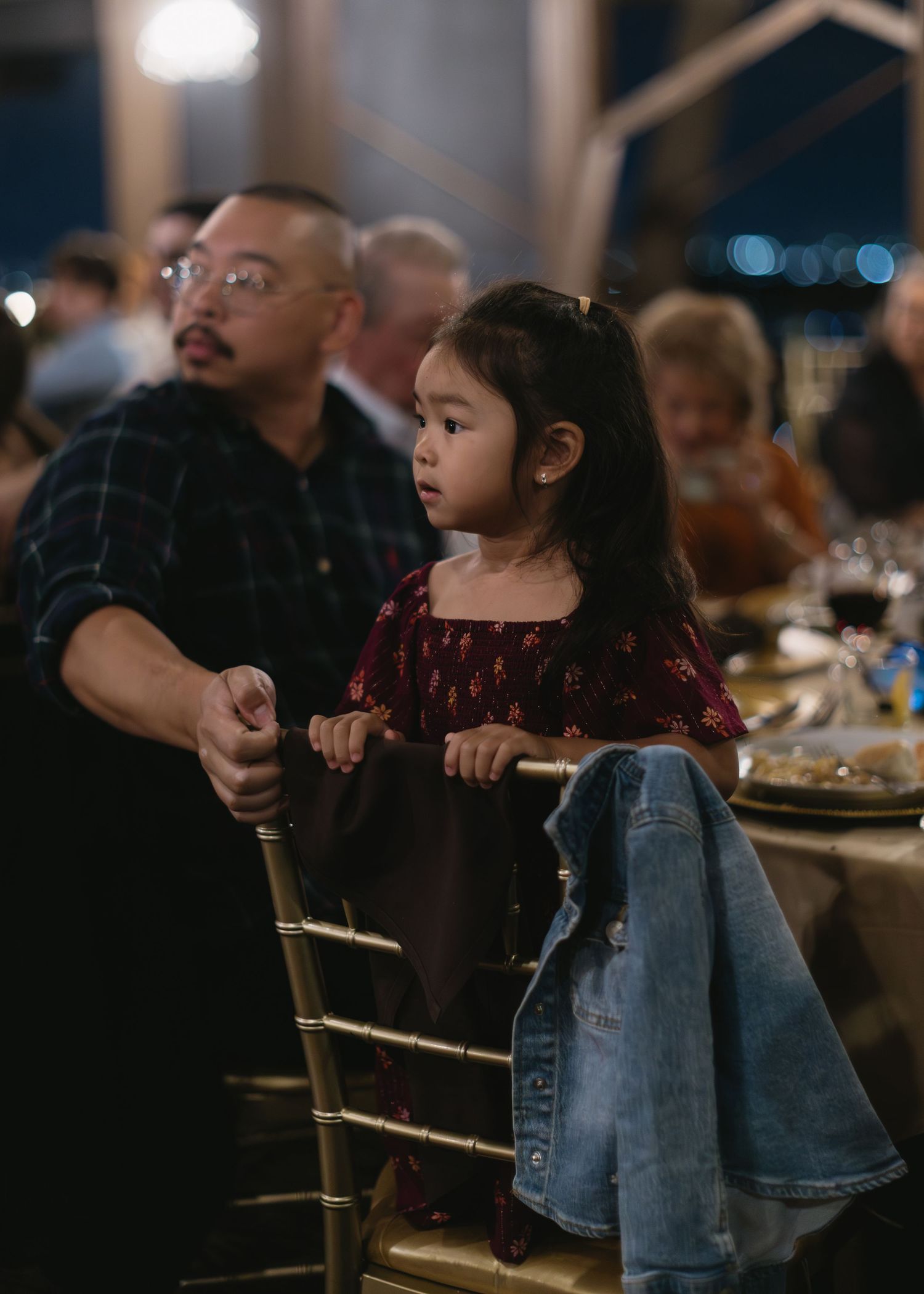 A young guest in denim and burgundy sits at an elegant reception table with gold chairs.