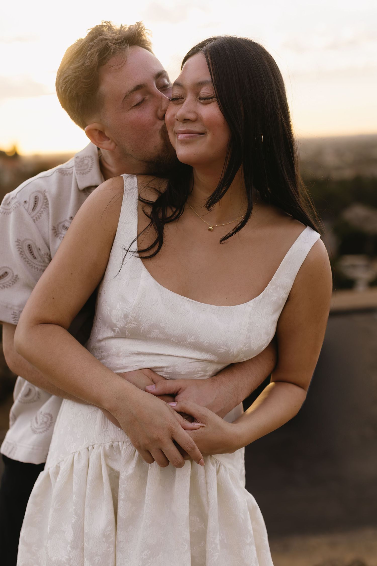 A romantic couple shares intimate moments during a sunset photoshoot, with the woman wearing a white dress.