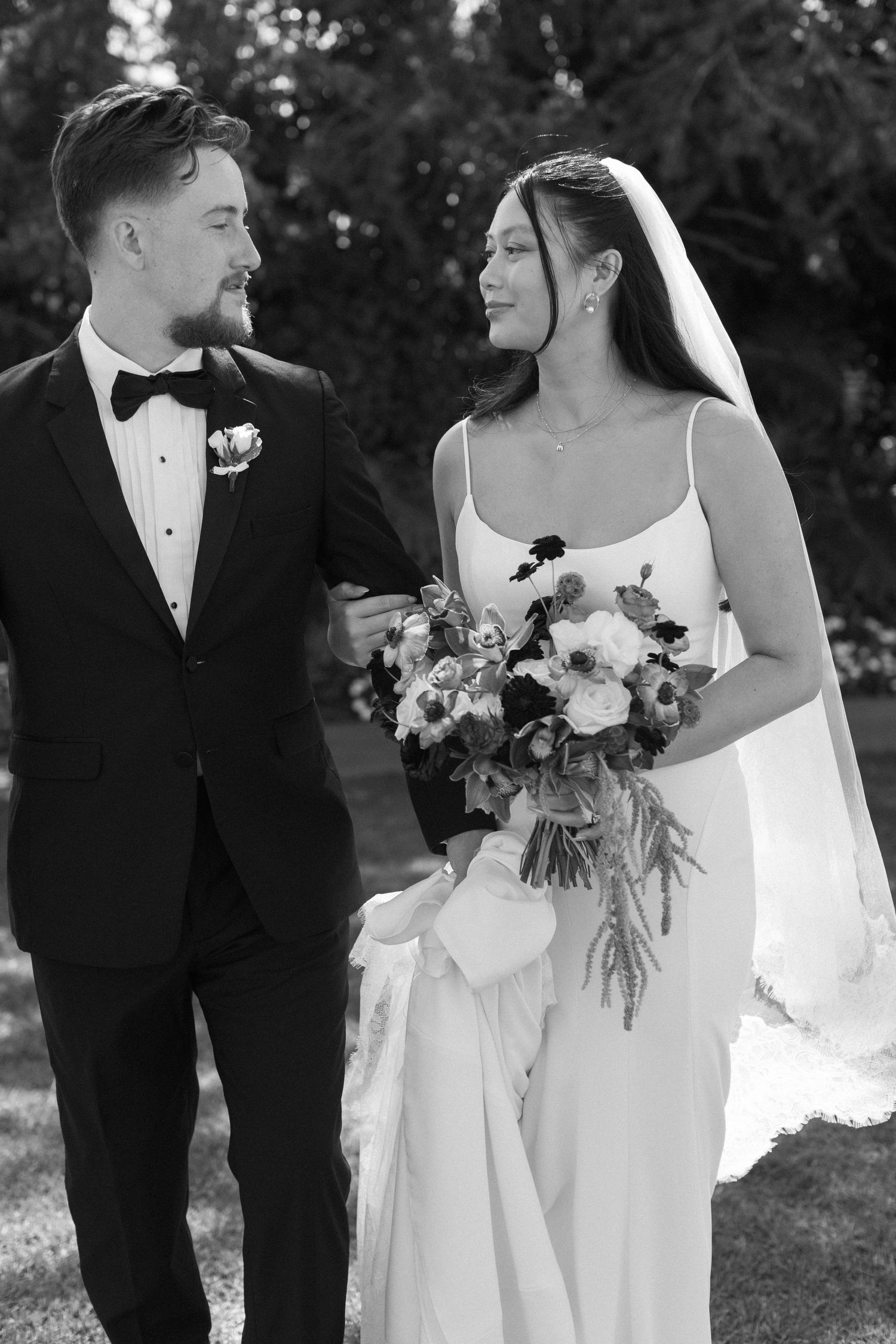 A black and white wedding portrait of a couple outdoors, with the bride holding a beautiful floral bouquet.