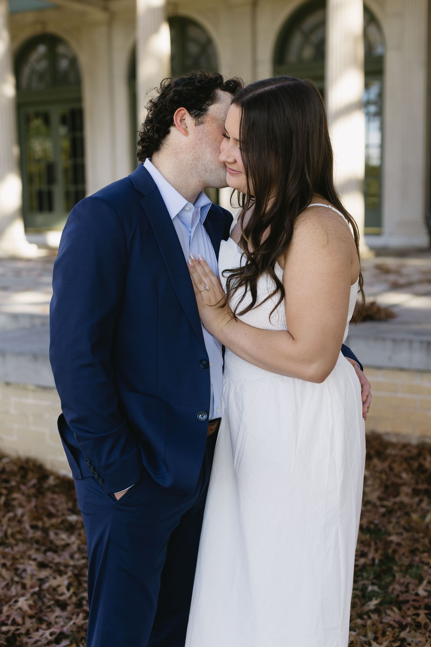 A couple shares a romantic kiss in front of a columned building with arched windows.