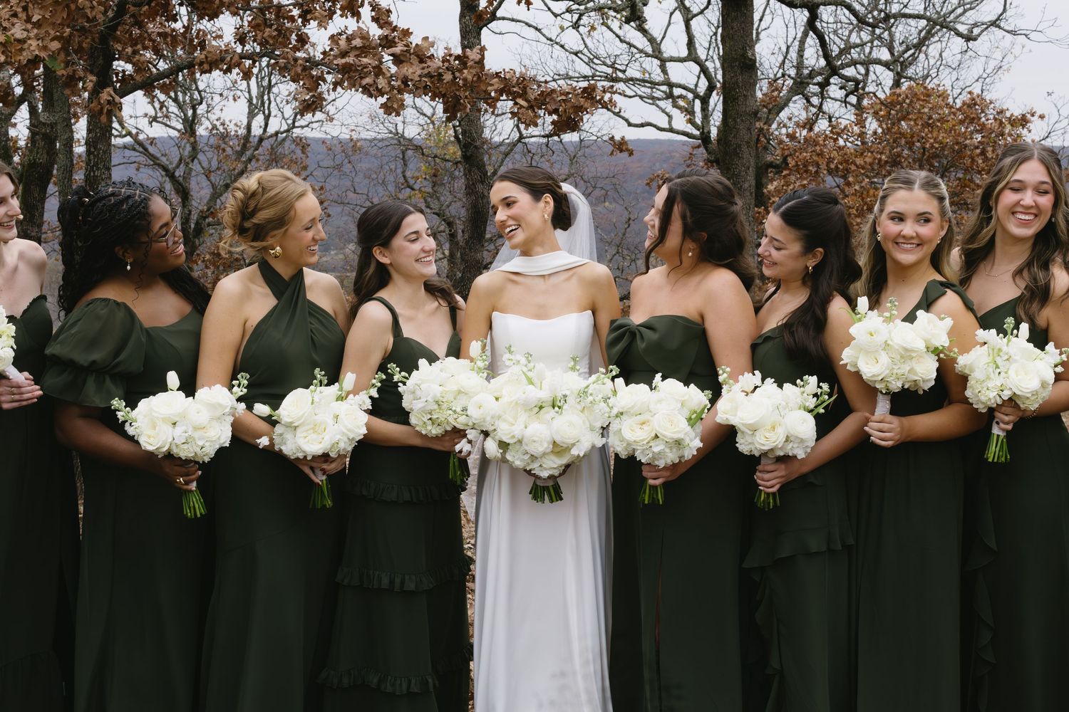Bridal party poses outdoors in autumn with bridesmaids in black dresses holding white floral bouquets.