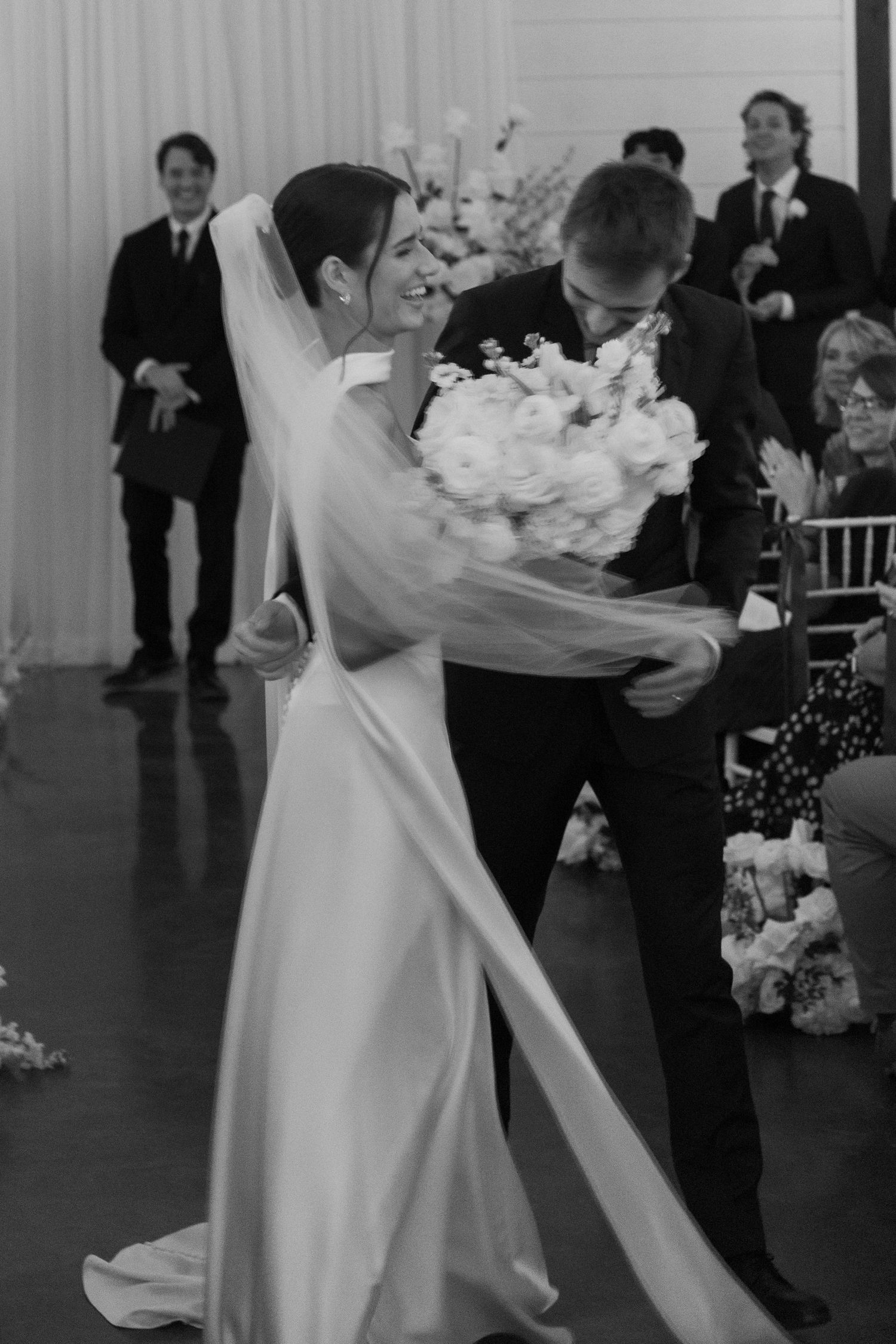 Black and white photo of couple dancing with white floral bouquets at indoor wedding reception.