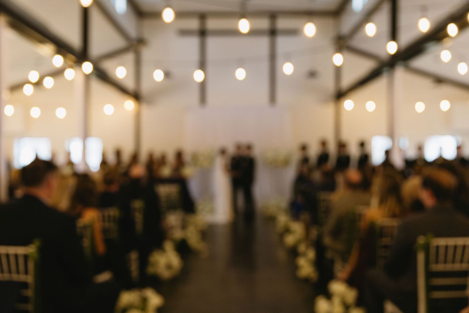 A blurred indoor wedding ceremony with string lights and guests seated in rows.
