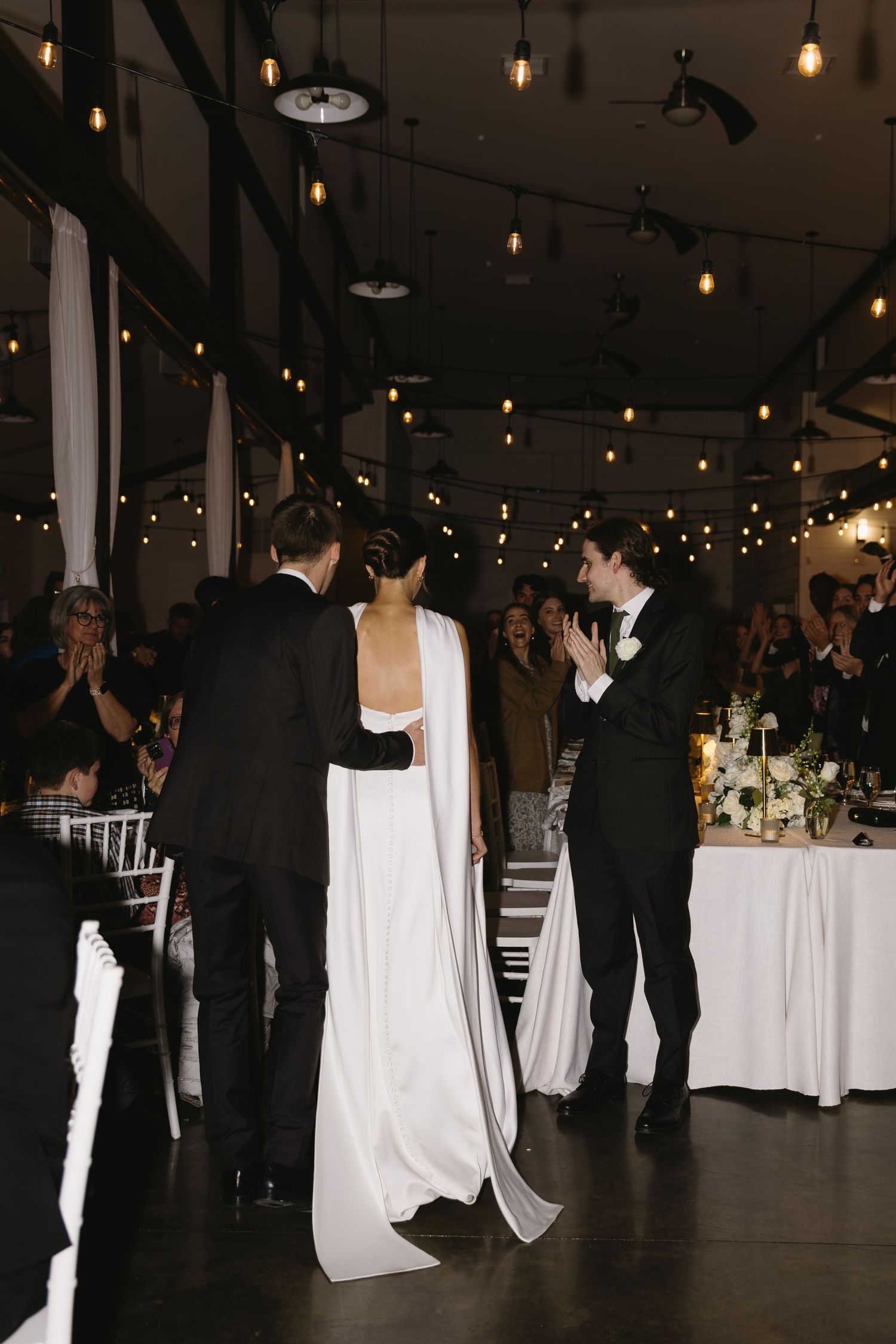 Wedding guests dancing in a dimly lit reception venue with string lights and white tablecloths.